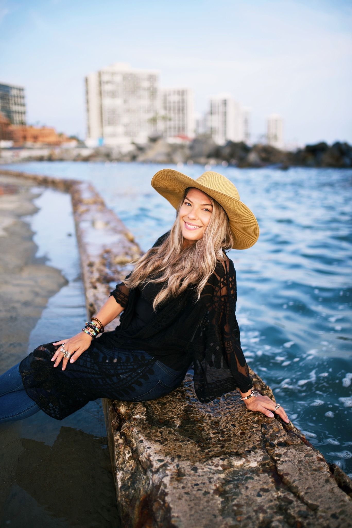 Seaside portrait session with black lace dress and wide-brim hat against blue ocean backdrop.