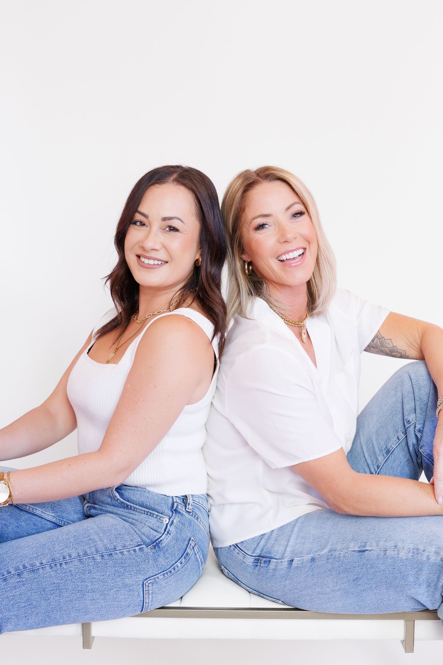 Two women in casual white tops and jeans sit back-to-back on white bench.