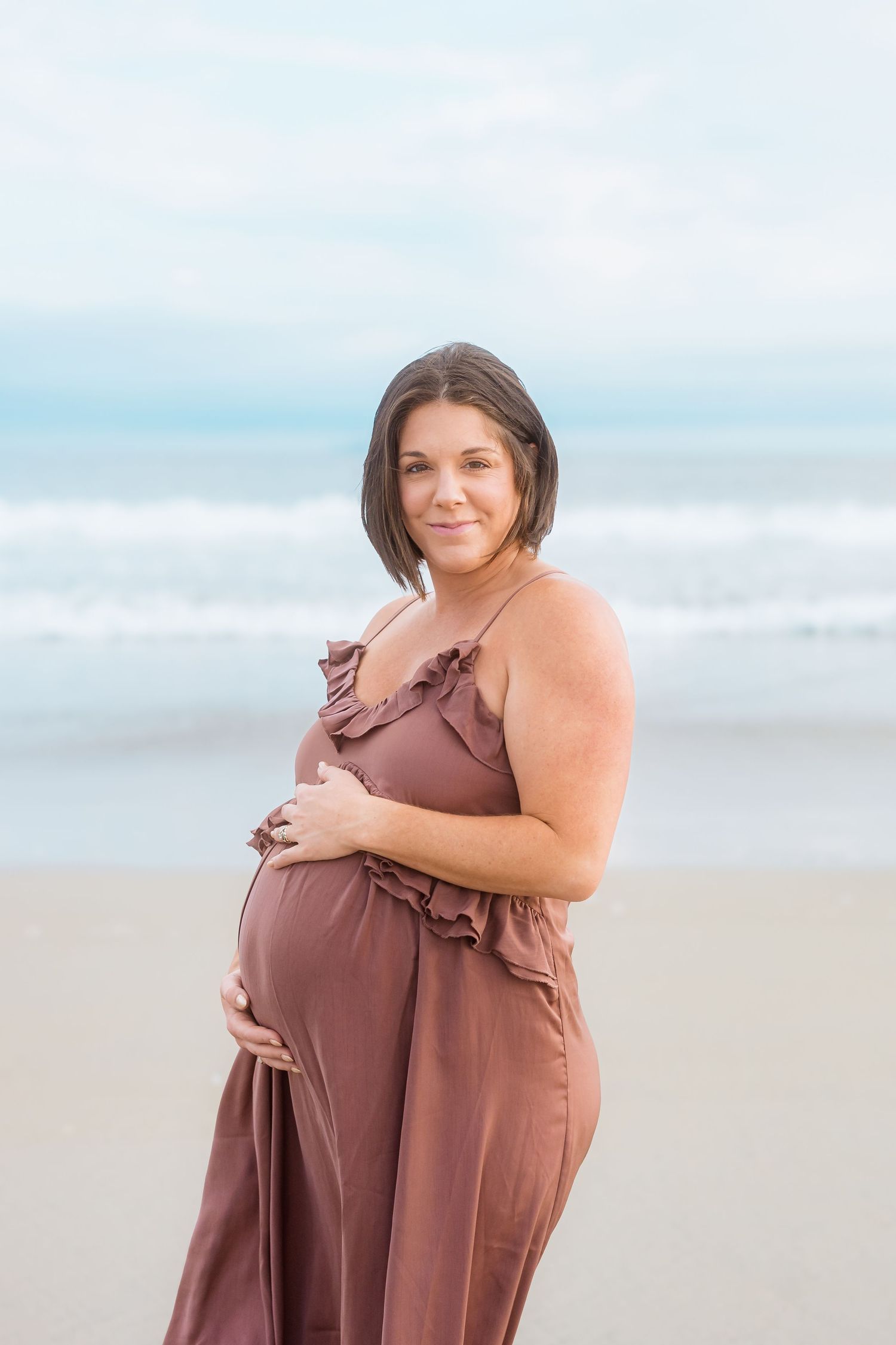 Expectant mother in mauve dress poses on beach during maternity photoshoot.