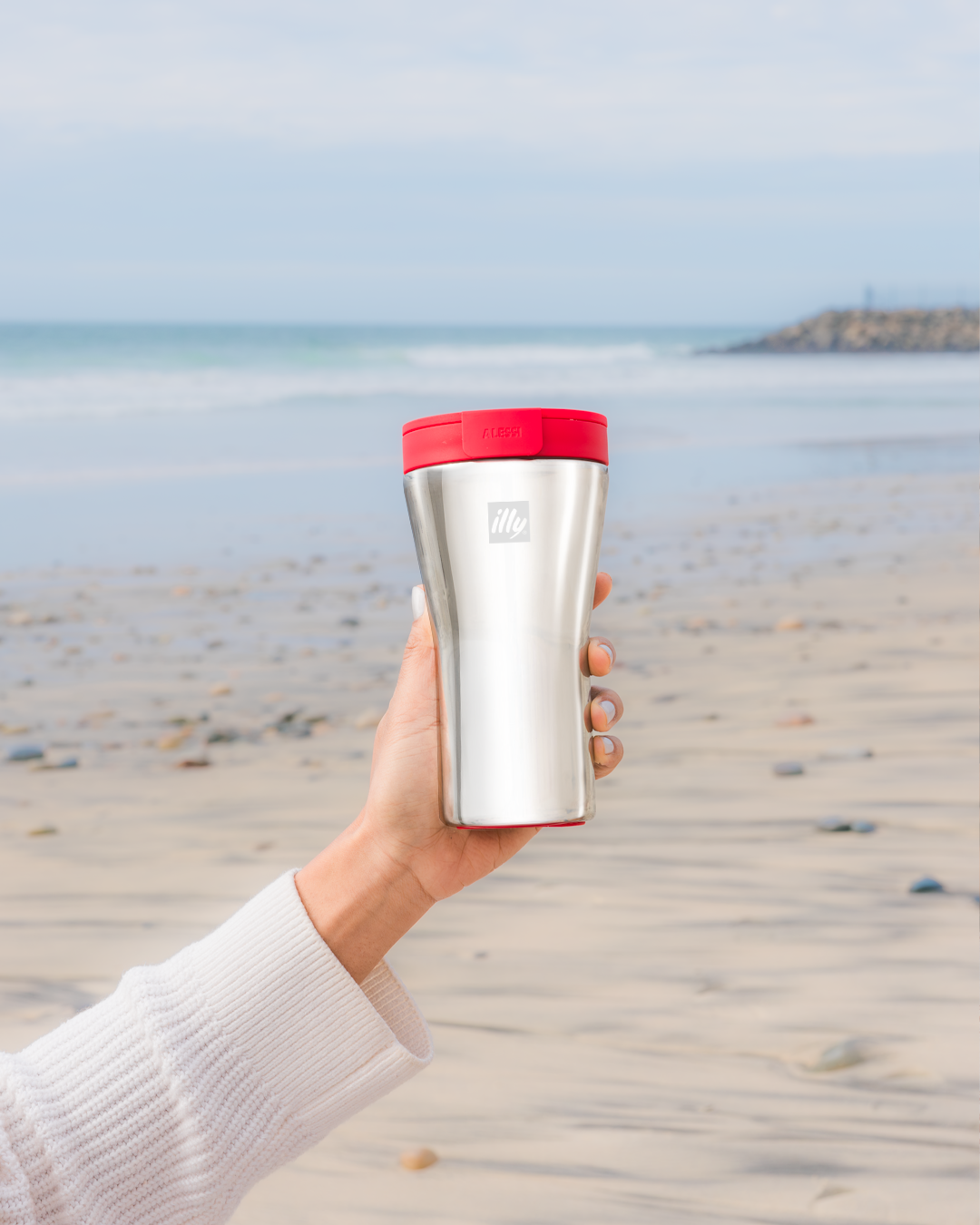 Silver travel mug with red lid held against beach backdrop.