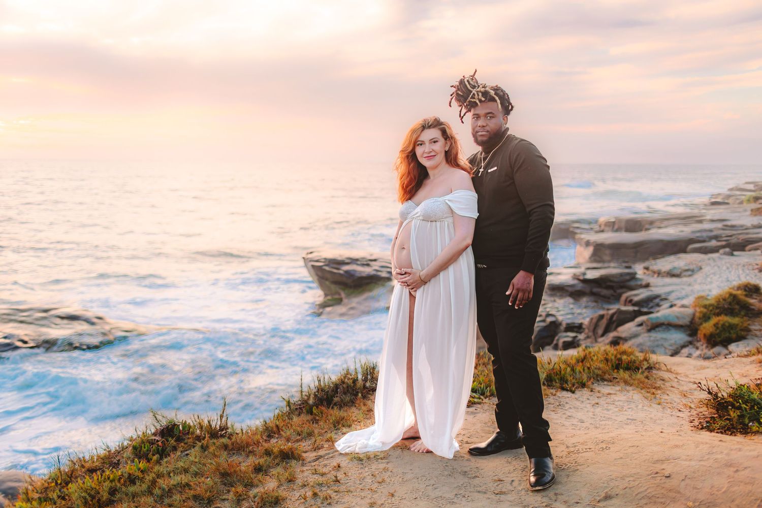 Couple in formal attire embraces on rocky coastal cliffs during sunset by the ocean.