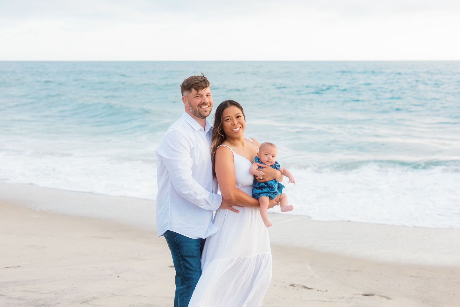 A sweet family moment captured at the beach with a baby wearing blue against turquoise ocean waves.