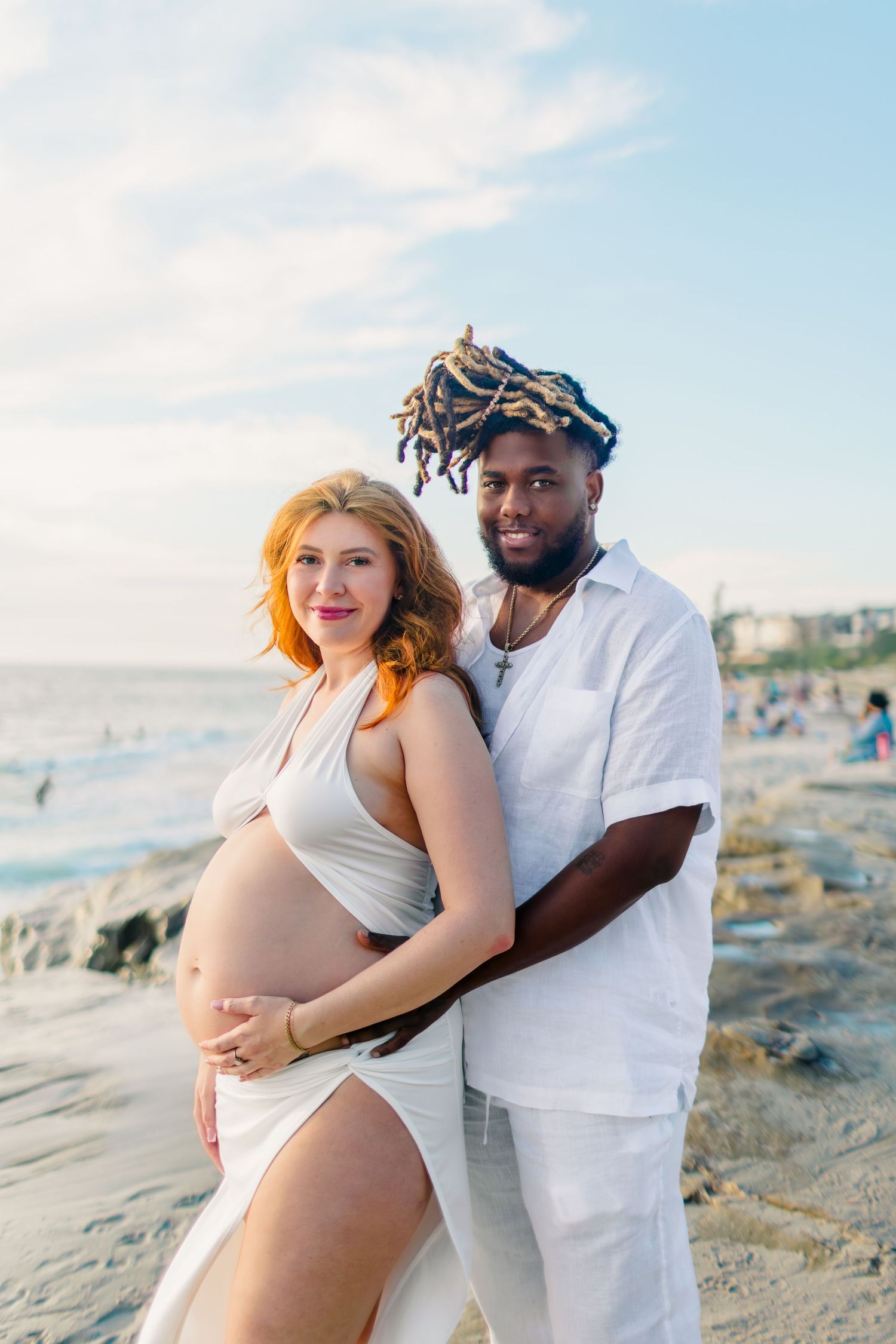 A couple embraces on a beach at sunset, with the pregnant woman wearing white and waves crashing in the background.