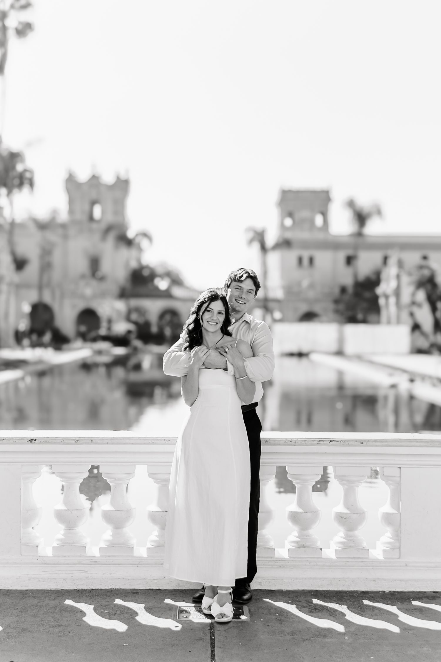 Elegant couple posing on a white balcony with castle-like architecture and palm trees in the background.