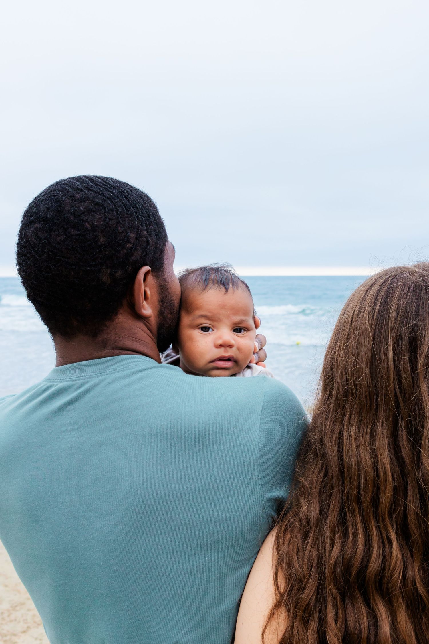 A couple holds their baby while standing on a beach with ocean waves in the background.
