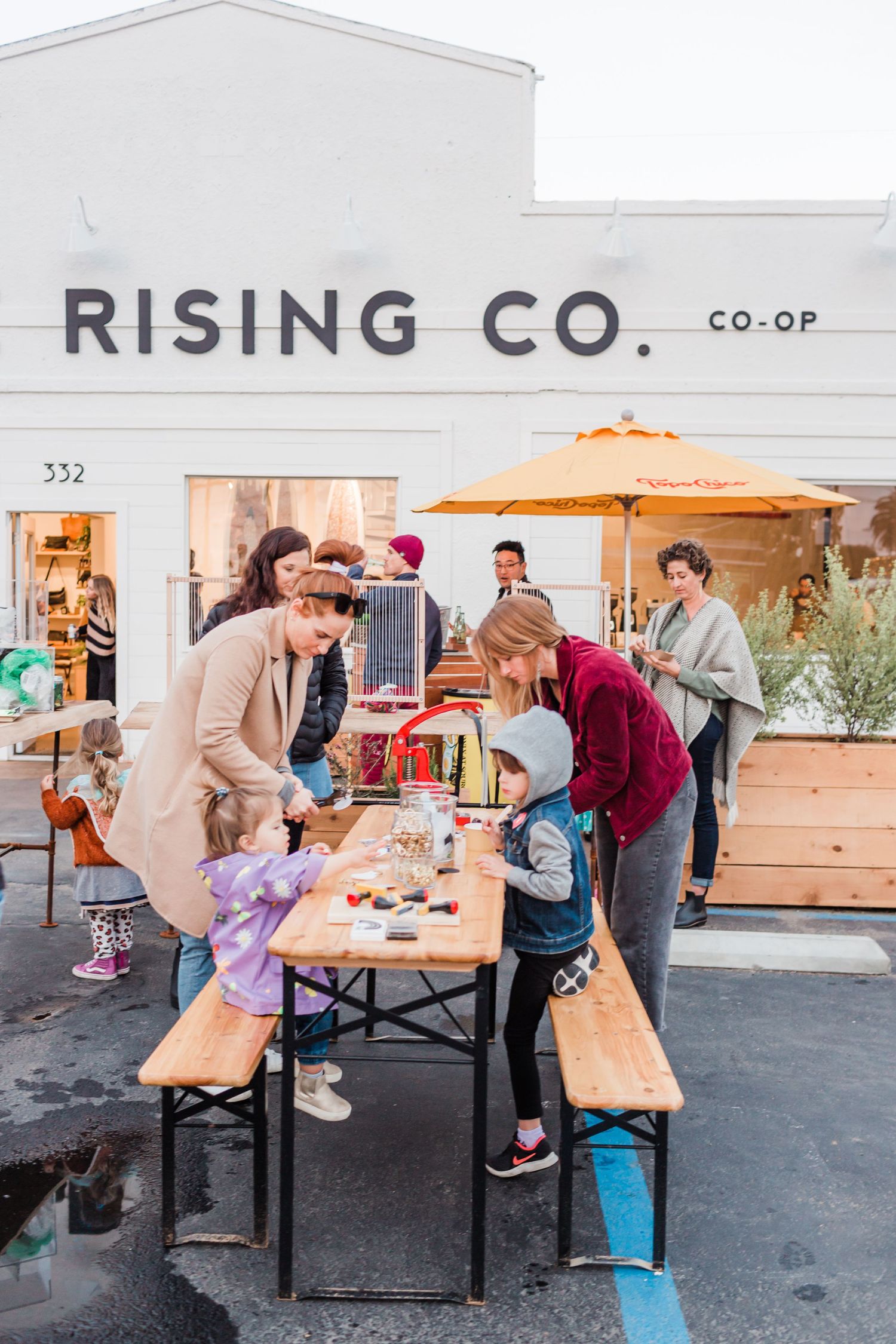 Outdoor gathering at Rising Co co-op with people sitting and standing at wooden picnic tables during daytime activities.