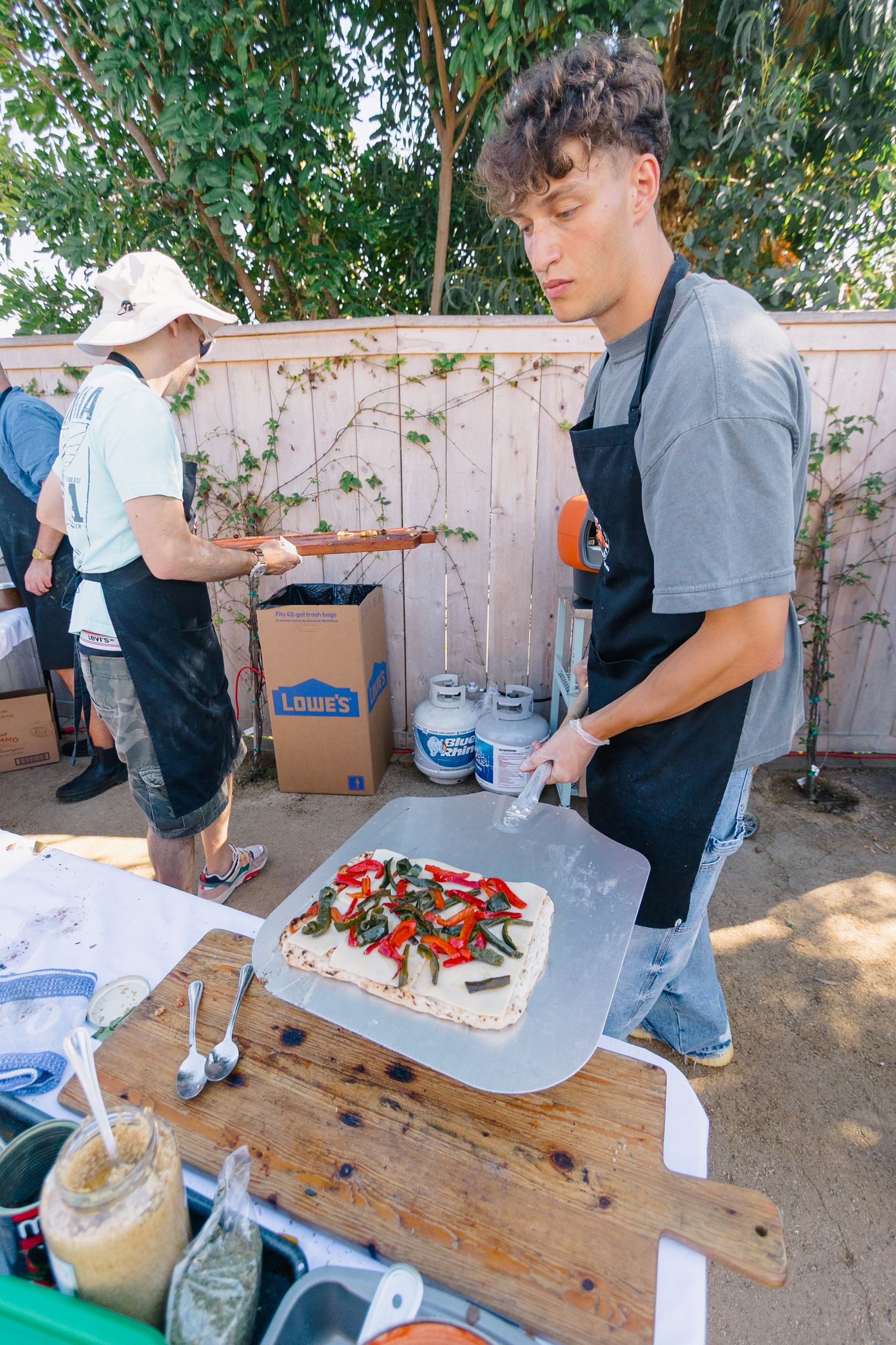 Outdoor buffet setup with people preparing food on wooden tables under trees.