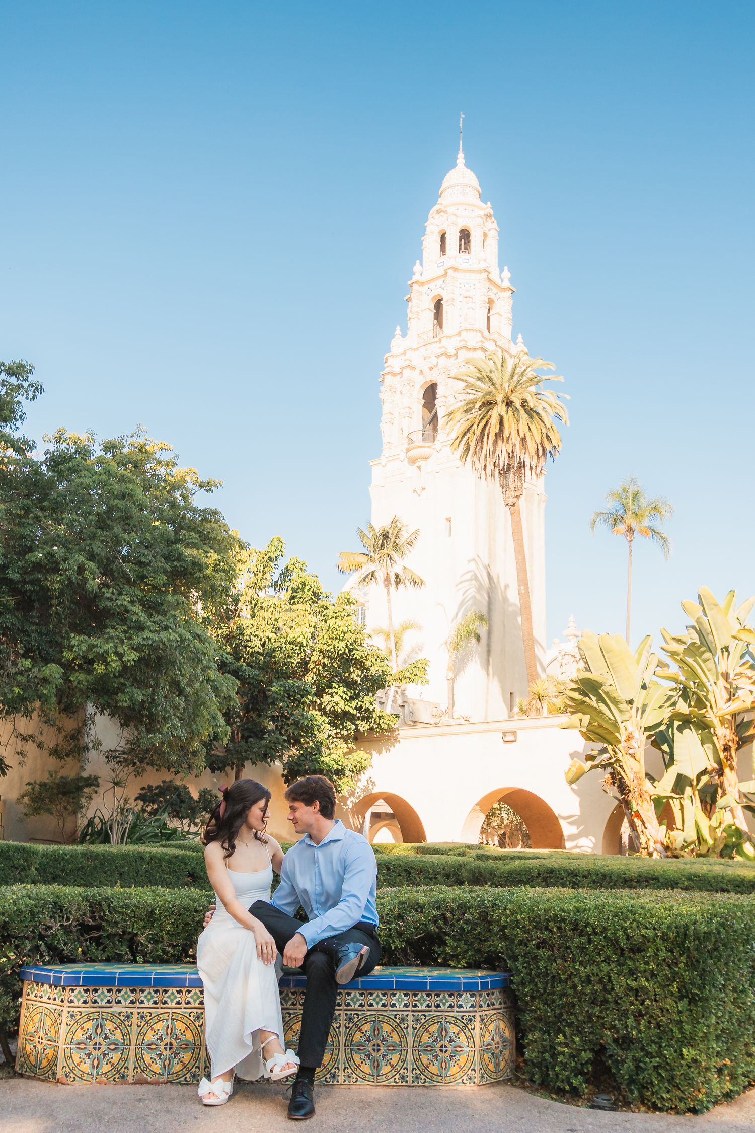 A couple sits on a decorative tiled bench in front of a white Spanish colonial bell tower with palm trees.