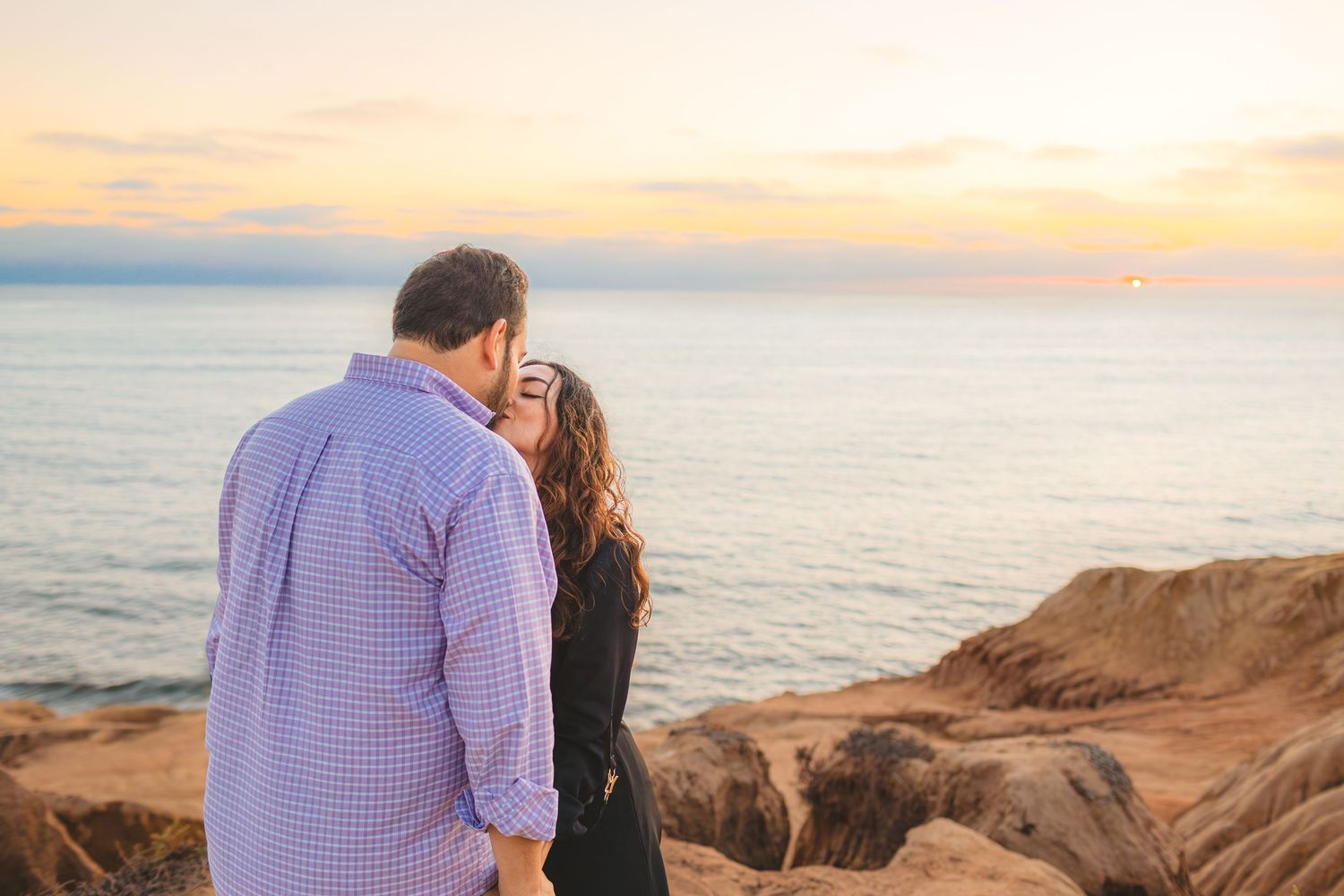 A couple shares a romantic moment on a rocky cliff overlooking the ocean at sunset.