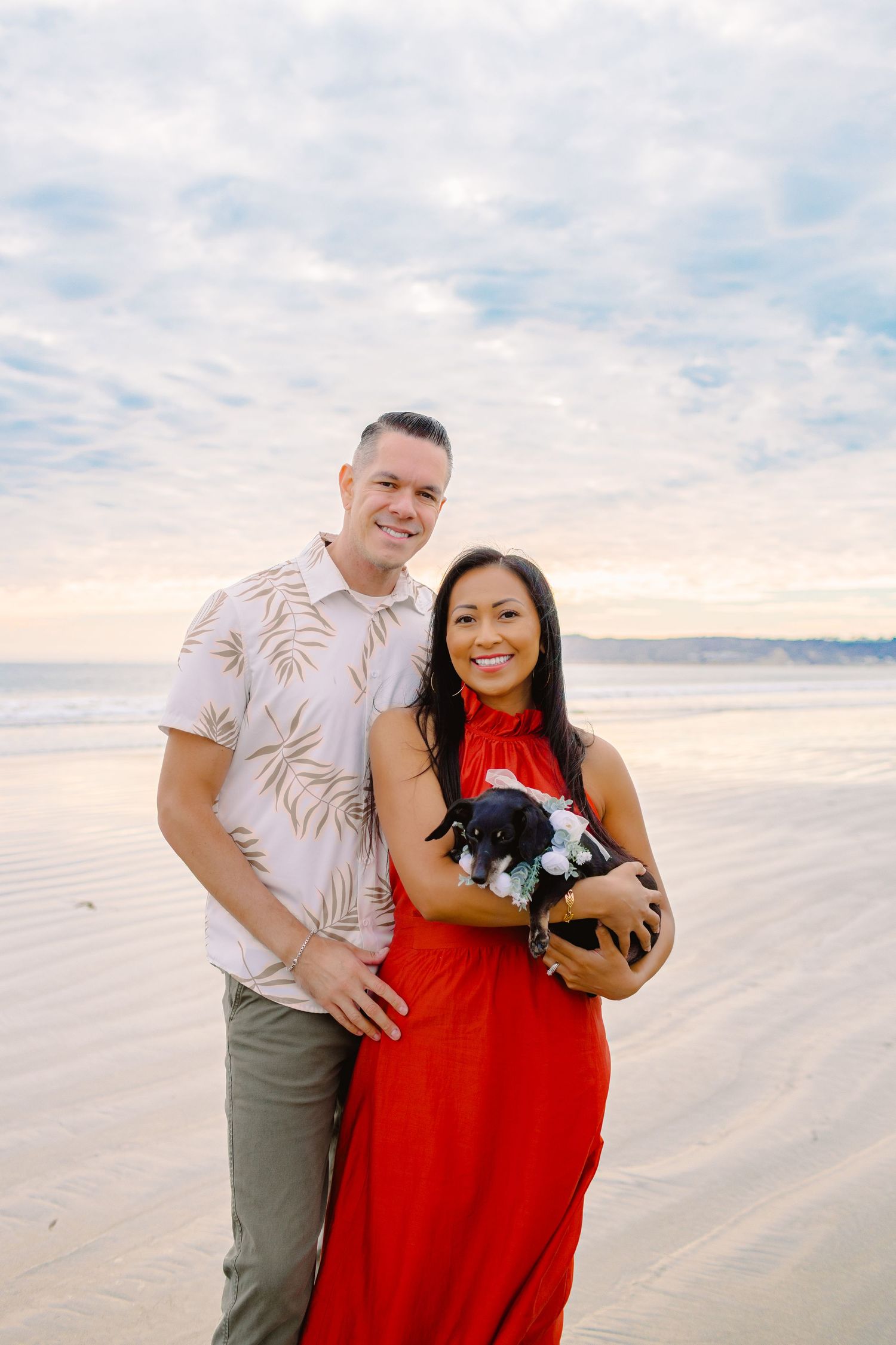 A couple and their small dog share a tender moment on a sandy beach at sunset.
