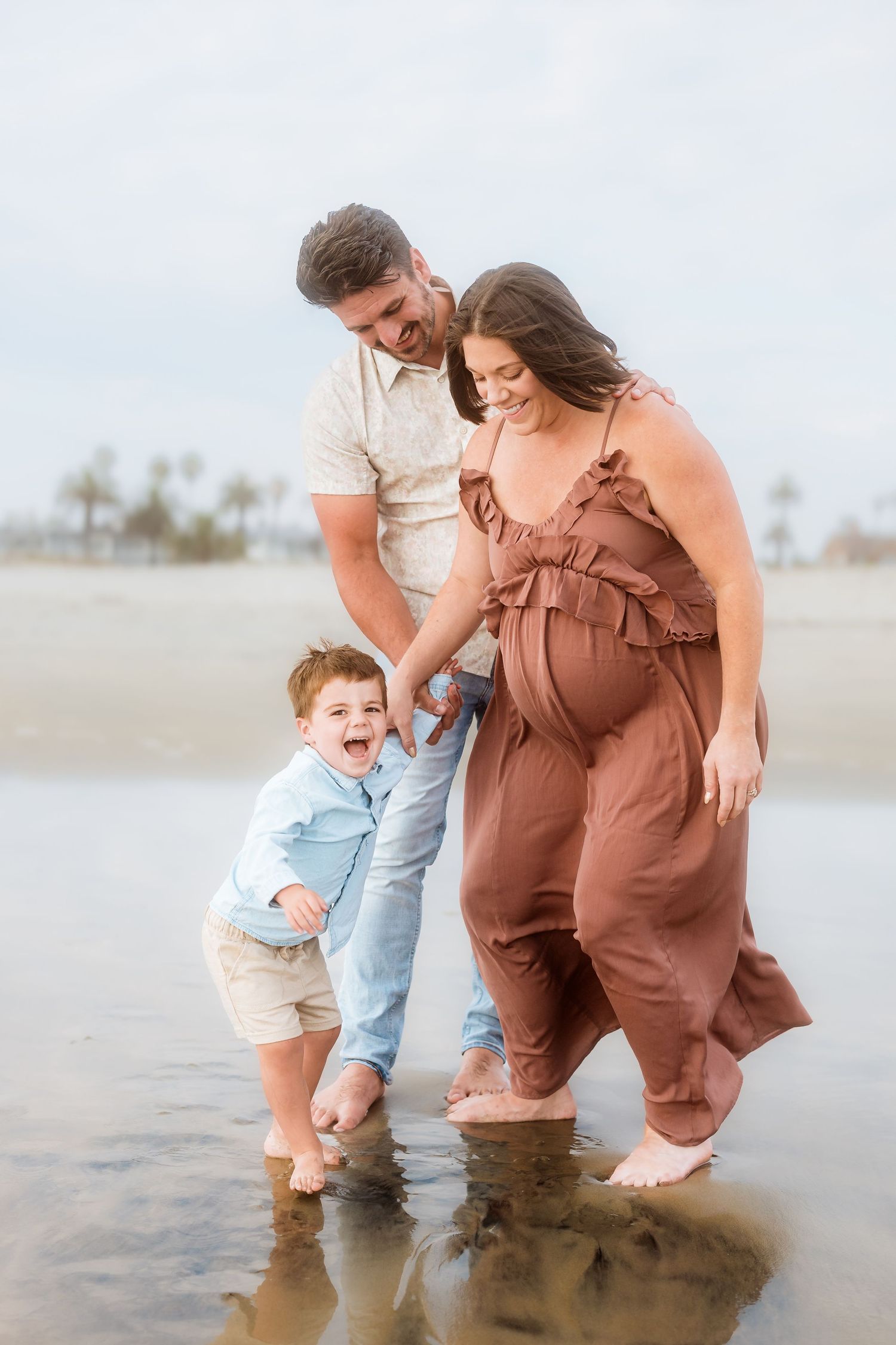 Family plays in shallow water at beach with toddler splashing between parents.