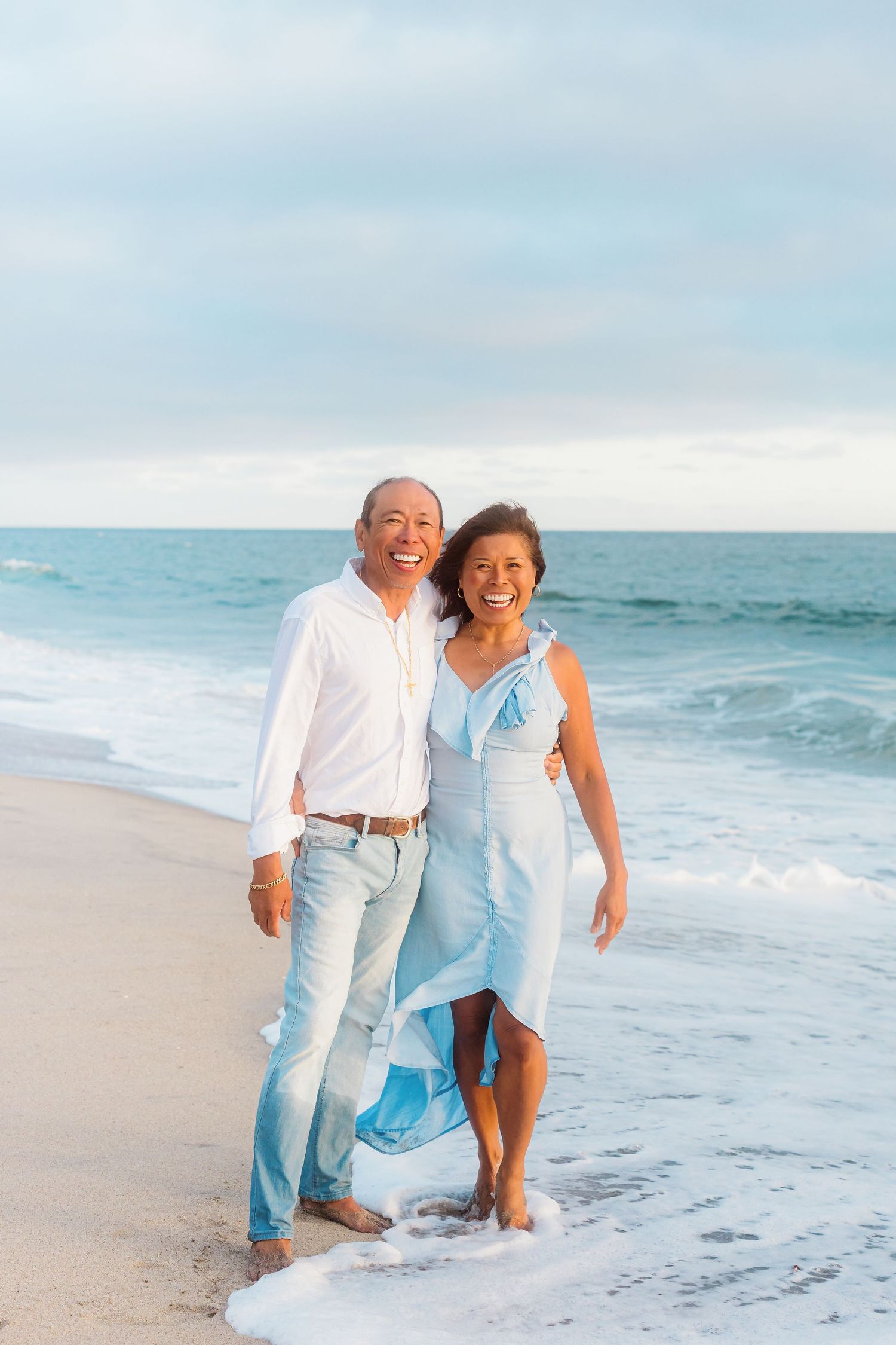 A couple stands together on the beach wearing light blue and white outfits.