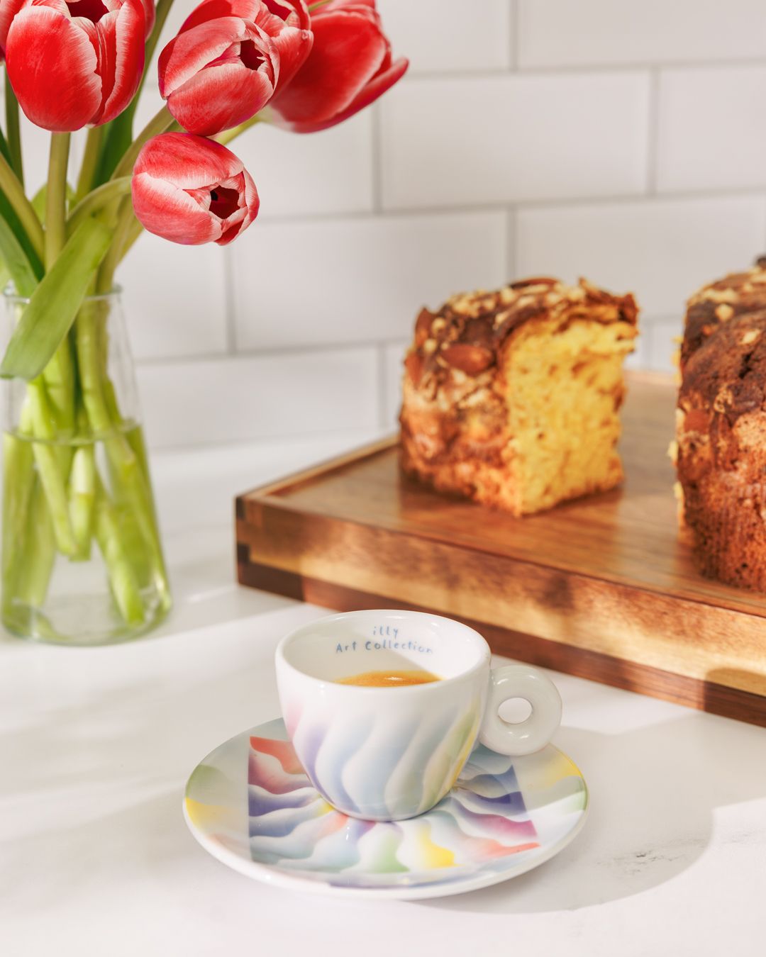 Red tulips in vase with coffee cup and baked goods on white counter.