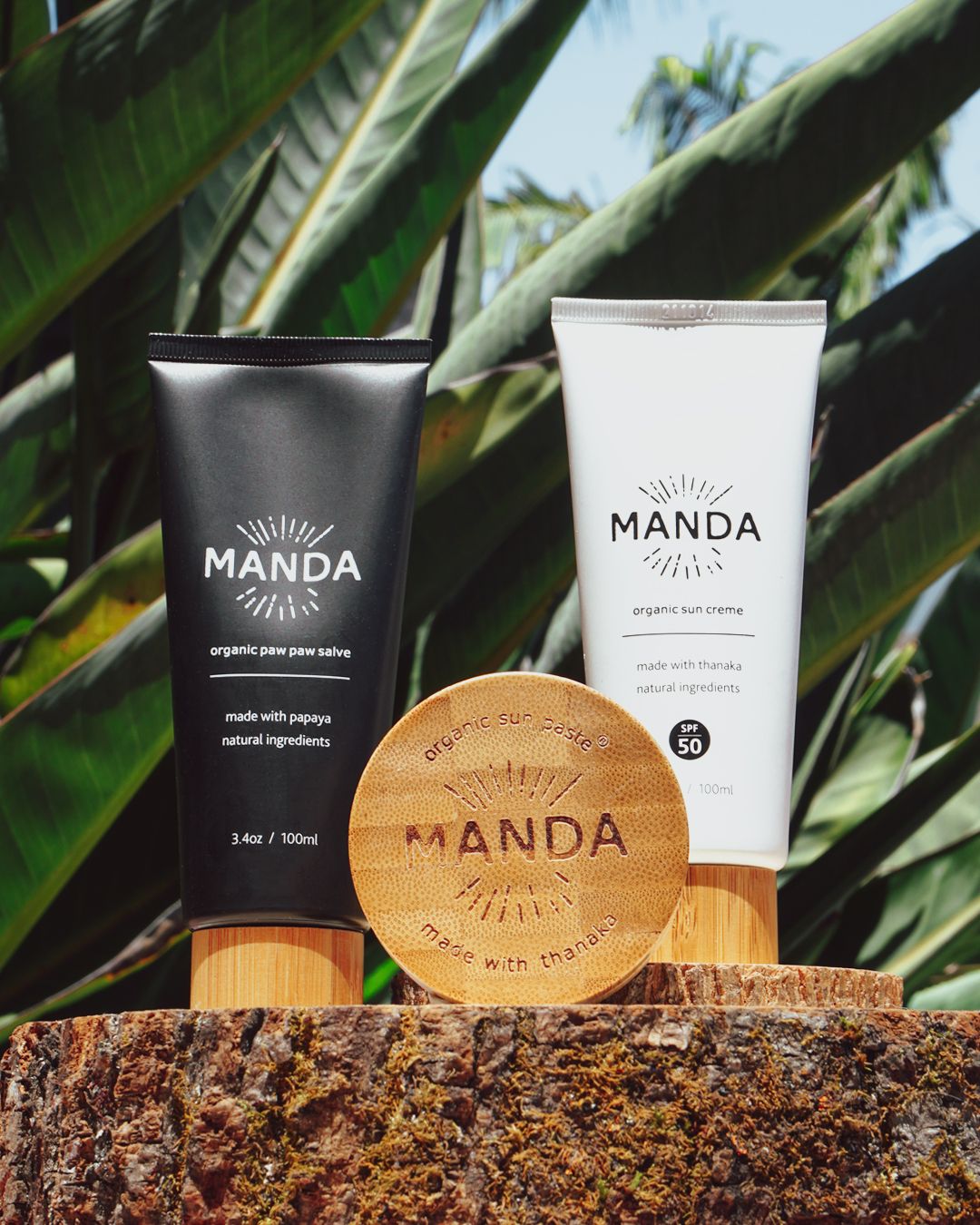 Black and white Manda skincare tubes with wooden compact displayed against tropical foliage.