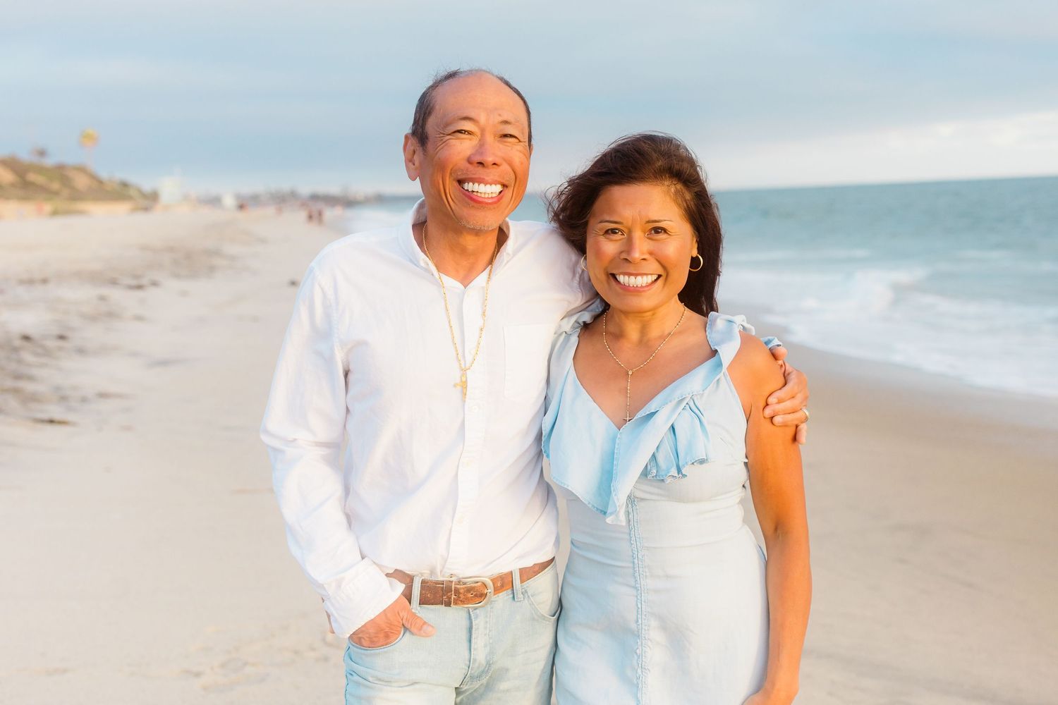 A happy couple poses on the beach wearing coordinated light blue and white outfits.