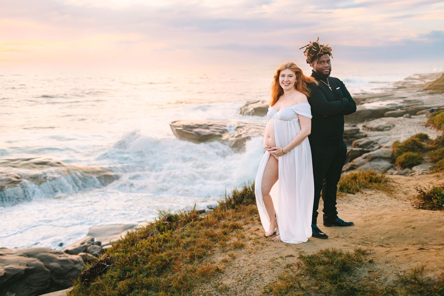Couple in formal attire embraces on rocky coastal cliffs at sunset with crashing waves in the background.