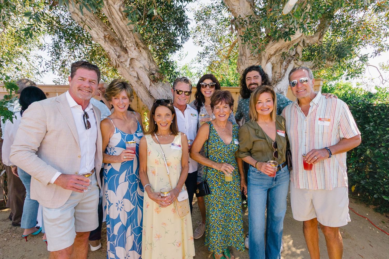 People gathered under trees at an outdoor party with white canopy tents.