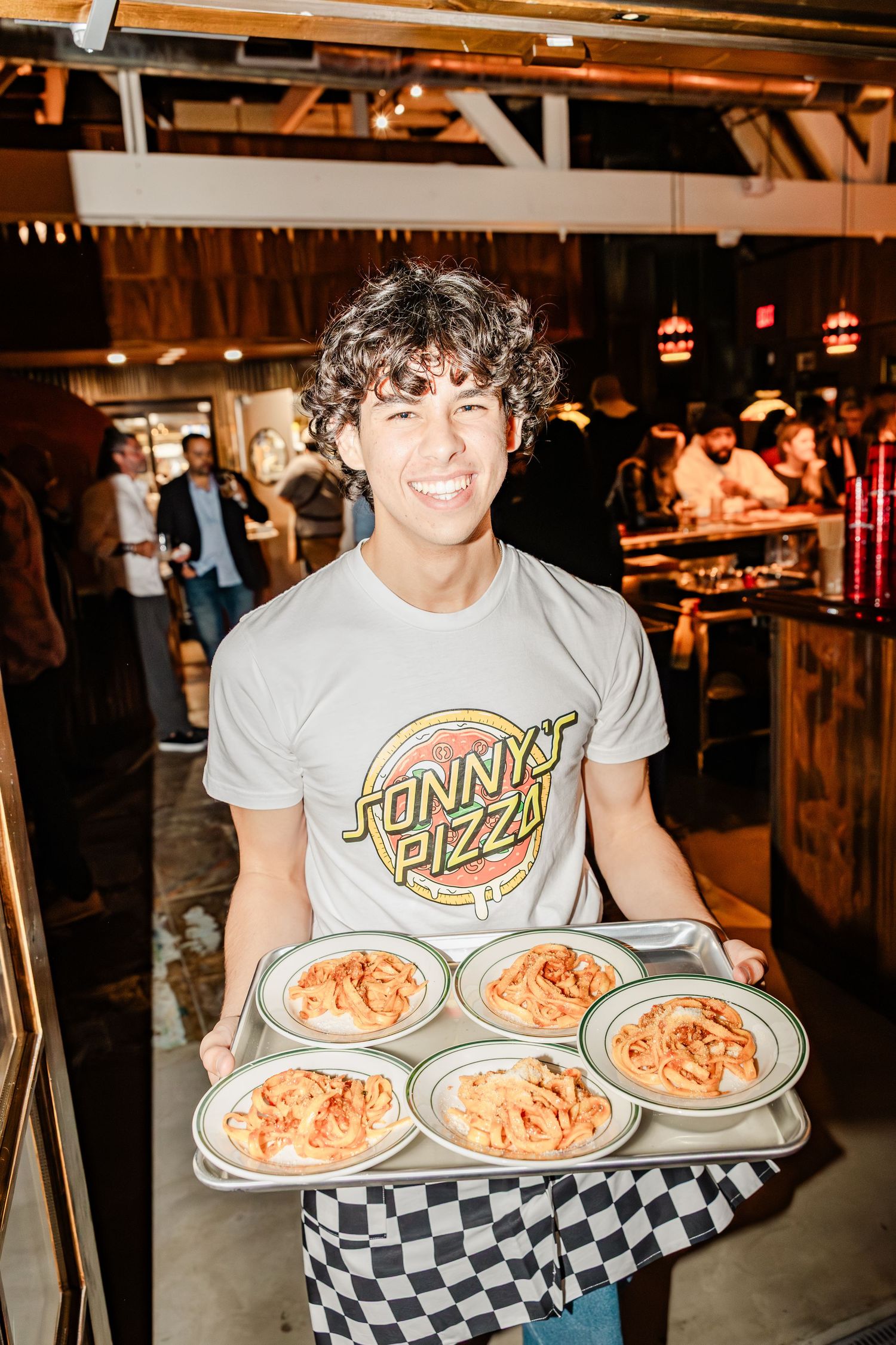 Server carrying multiple plates of pasta on trays through a dimly lit restaurant.
