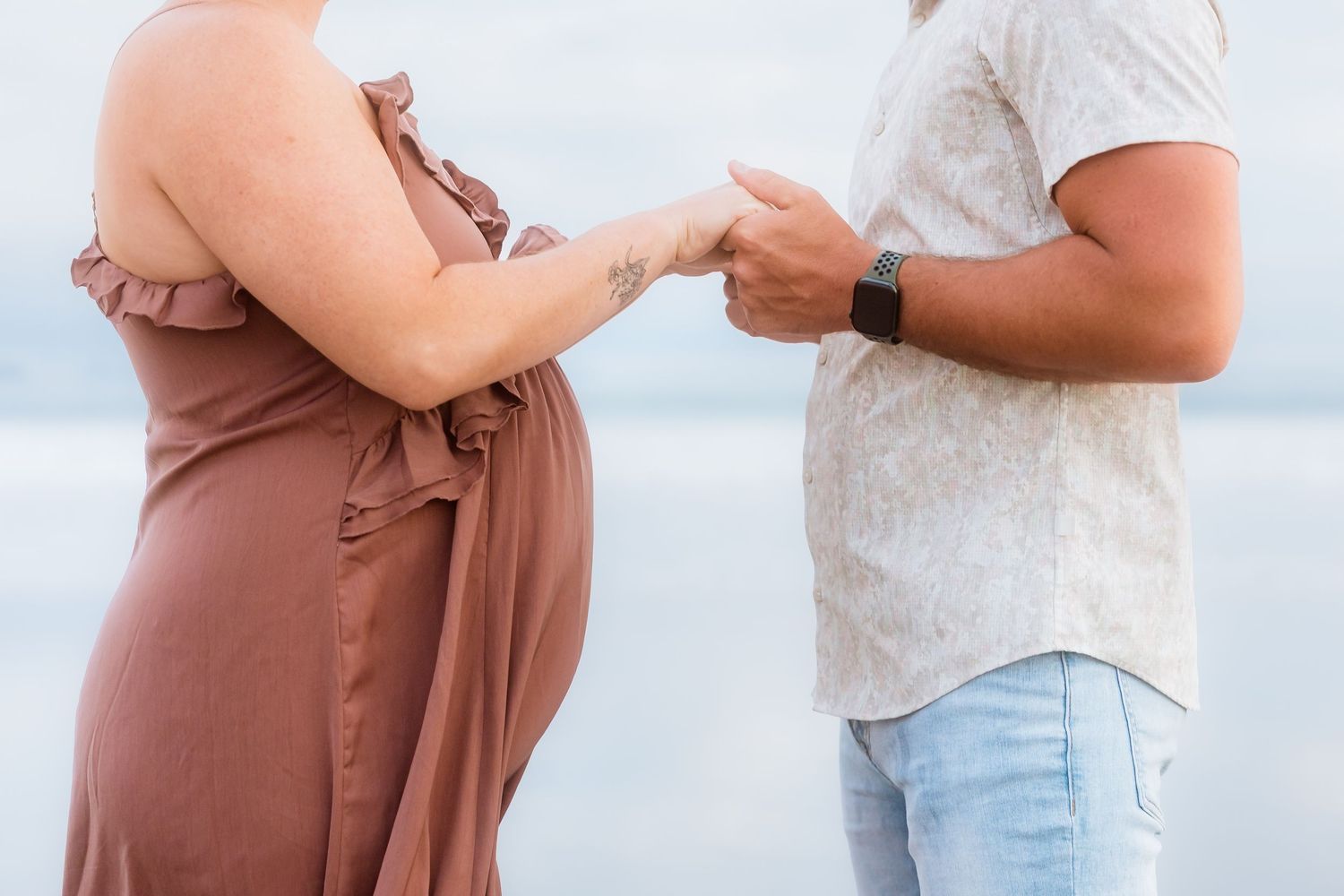 Close up of couple holding hands on beach with woman in mauve dress showing baby bump.