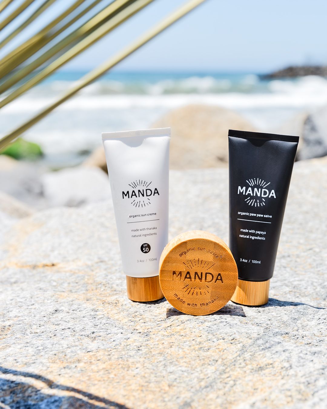 Manda skincare collection featuring black and white tubes with wooden compact on beach rocks.