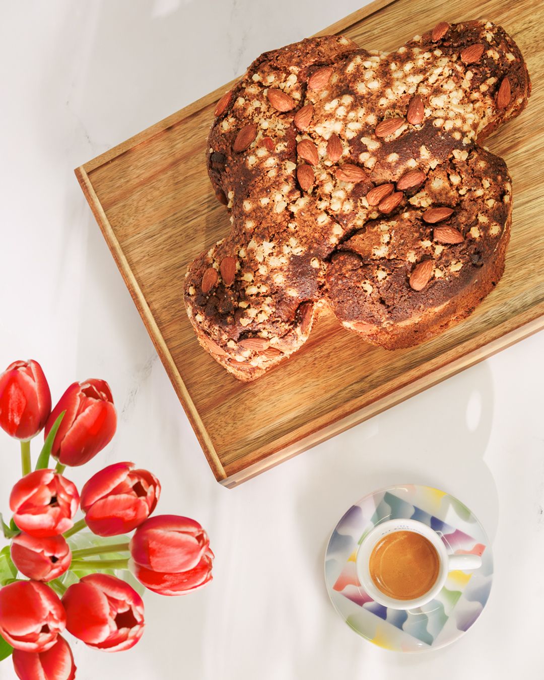 Rustic bread loaf on wooden board with red tulips and coffee cup.