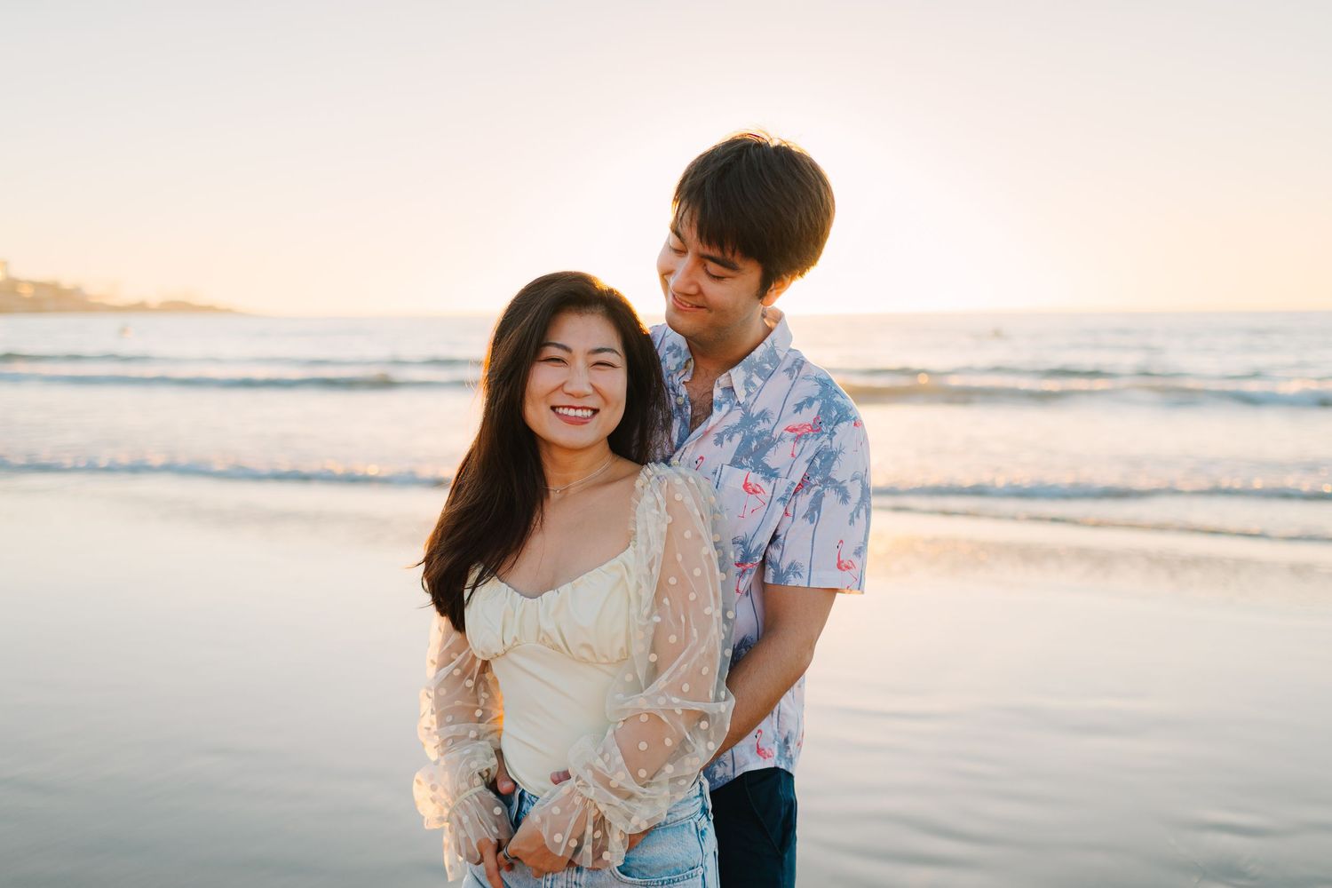 A couple shares a romantic moment together on the beach at sunset while waves roll in behind them.