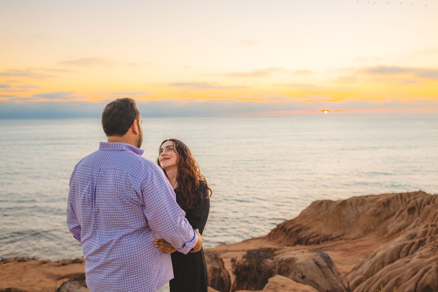 A couple embraces on a scenic cliff overlooking the ocean during a golden sunset.