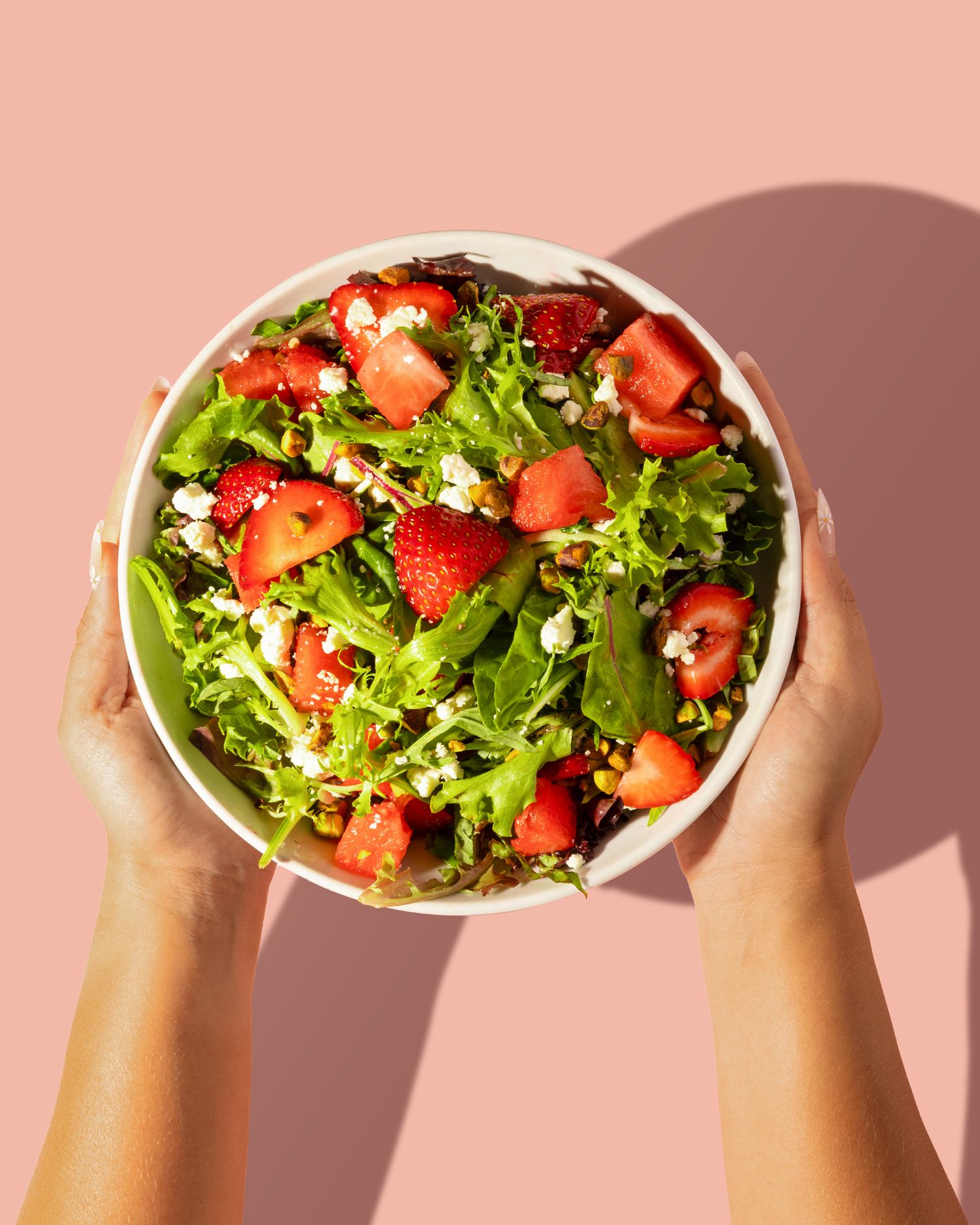 Hands holding bowls of fresh strawberry and green salad against a pink background.