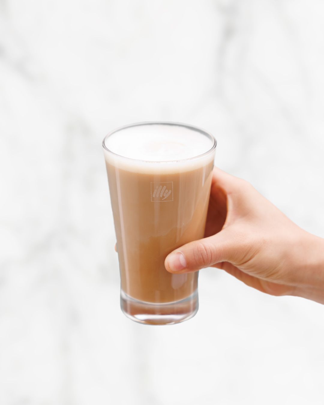 Hand holding glass of iced latte with foam against white marble background.