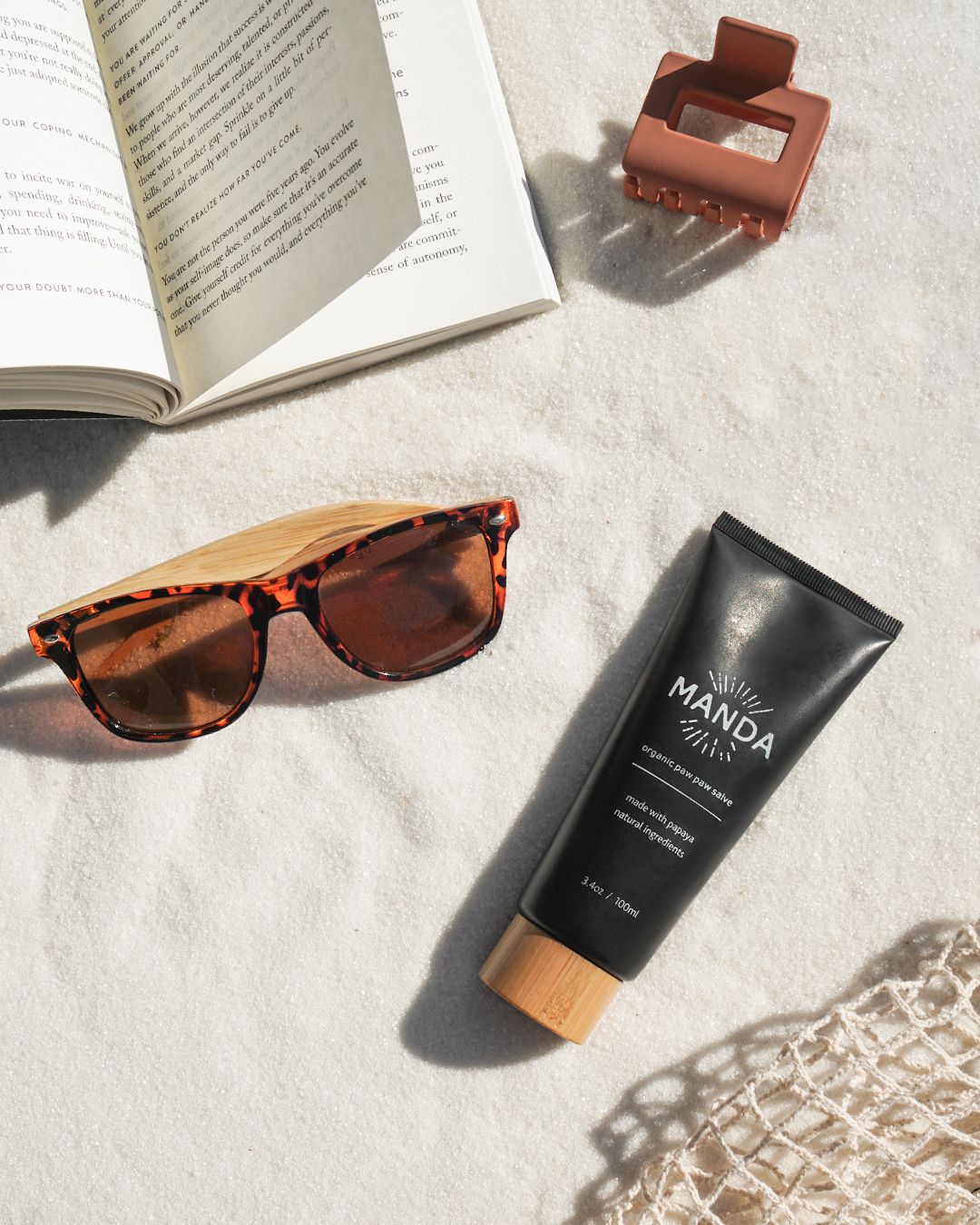 Lifestyle flatlay of sunglasses, book, and Manda skincare products on concrete surface.