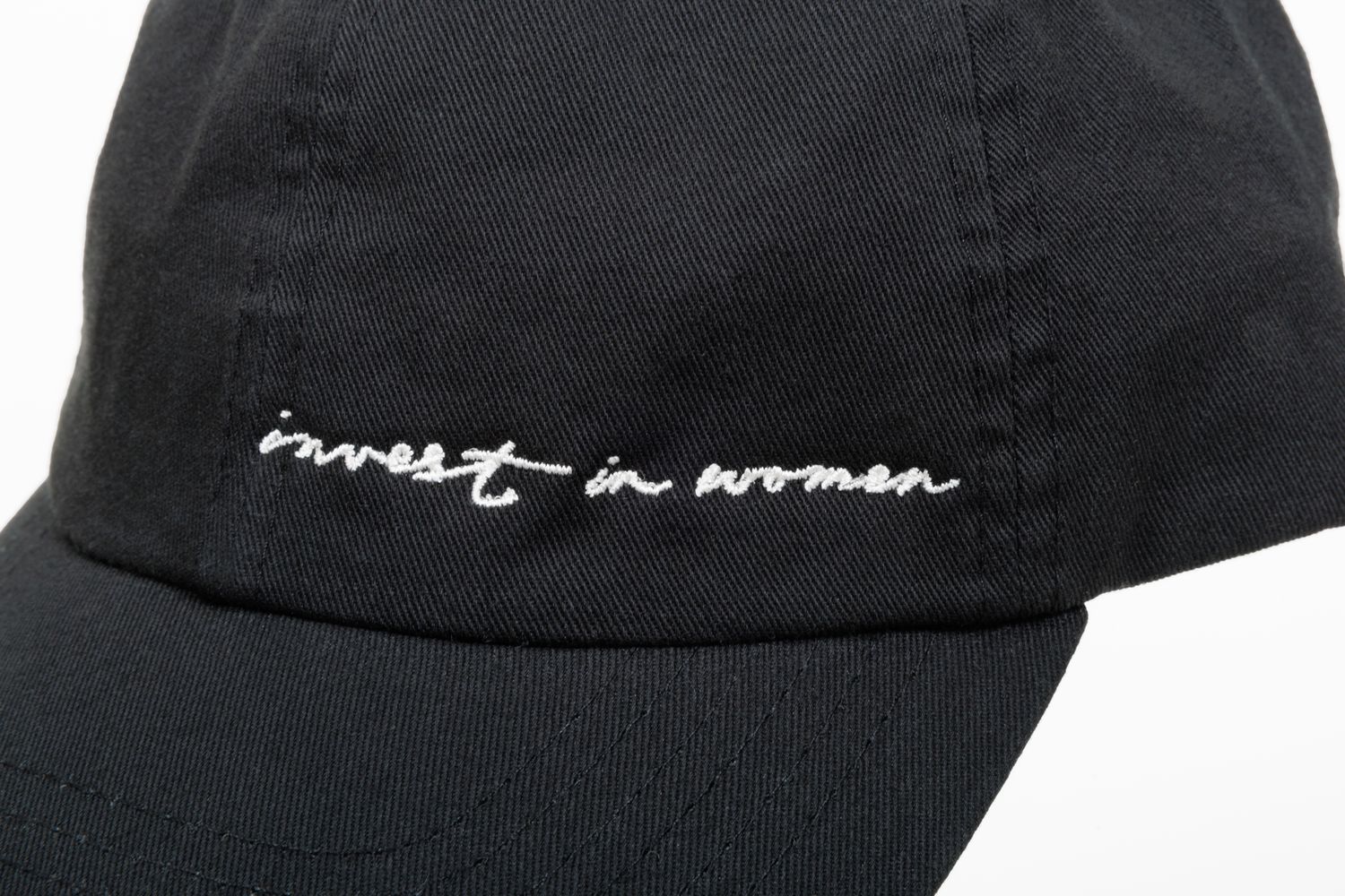Close-up of black cap showing 'invest in women' embroidered text detail.