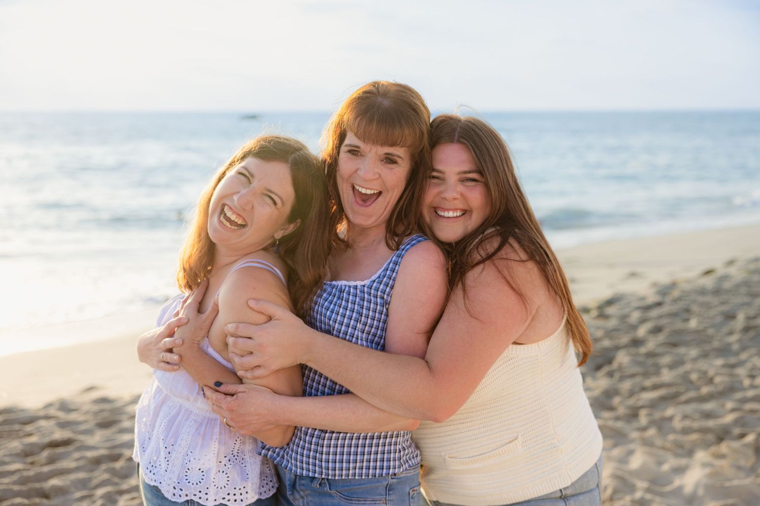 Three people share joyful embraces while posing together on a sunny beach with ocean waves in the background.
