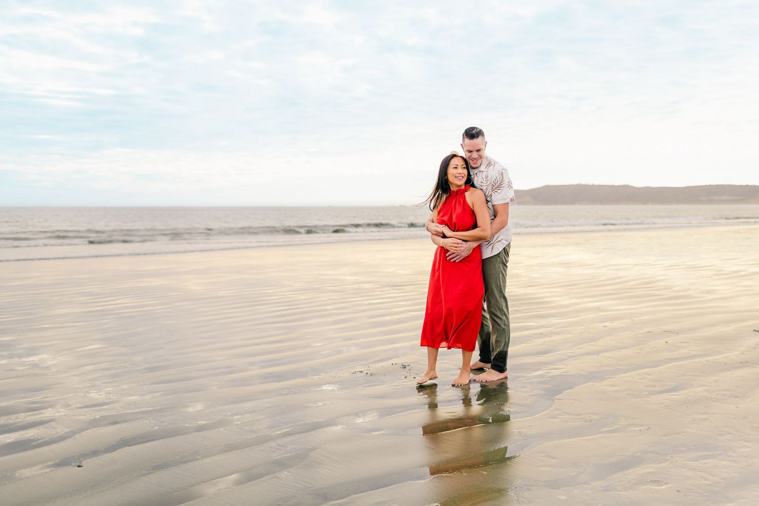 A couple embraces on a vast empty beach with a red dress flowing in the coastal breeze.