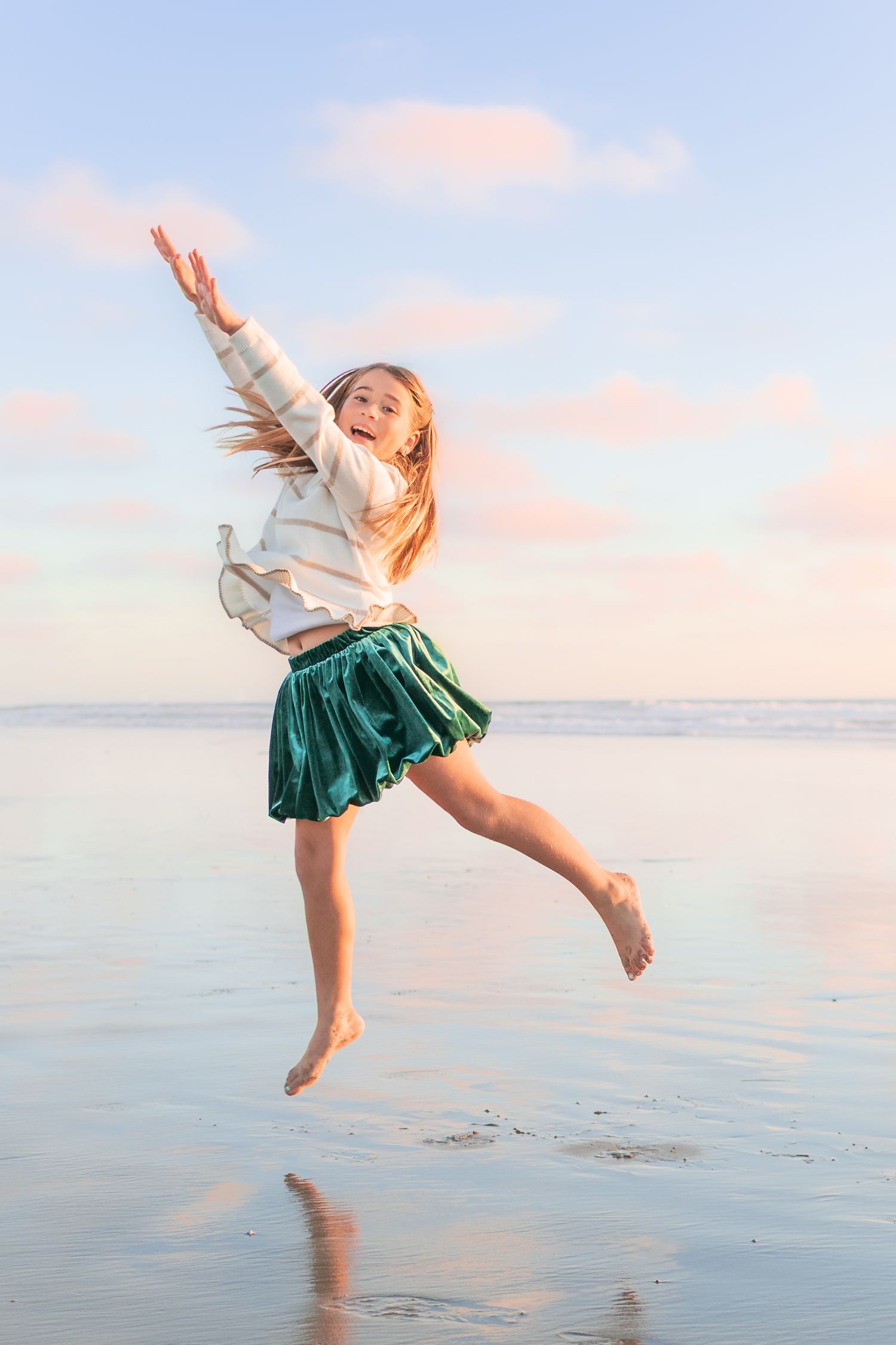 Woman in green skirt and white top dances joyfully on beach at sunset with pink sky in background.