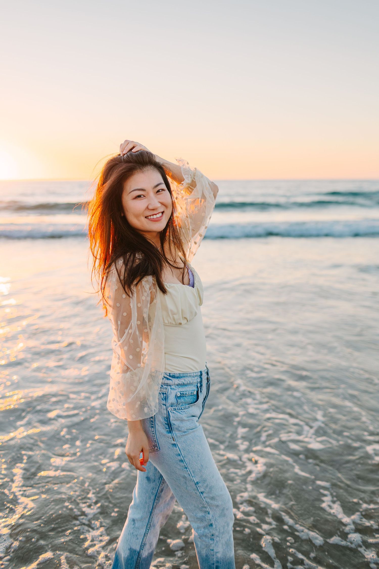 Young woman in lace top and jeans poses playfully on beach during golden hour.