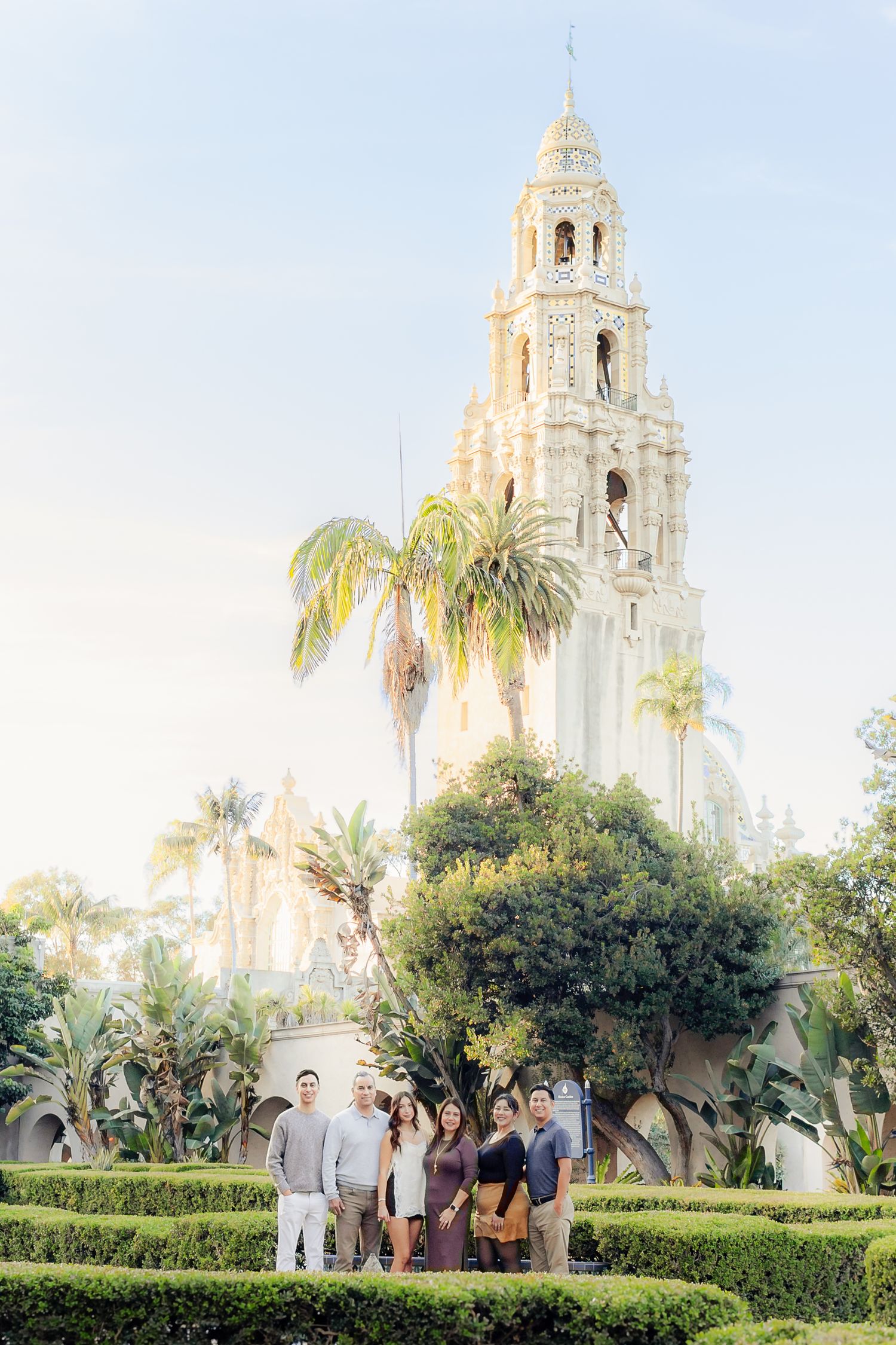 A group stands in a manicured garden with a Spanish colonial bell tower and palm trees rising behind them.