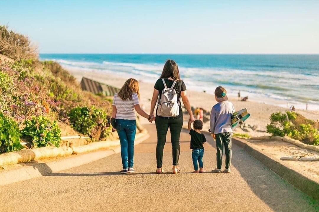 People walking together down coastal path with ocean view and pink flowers along California beach on sunny day.