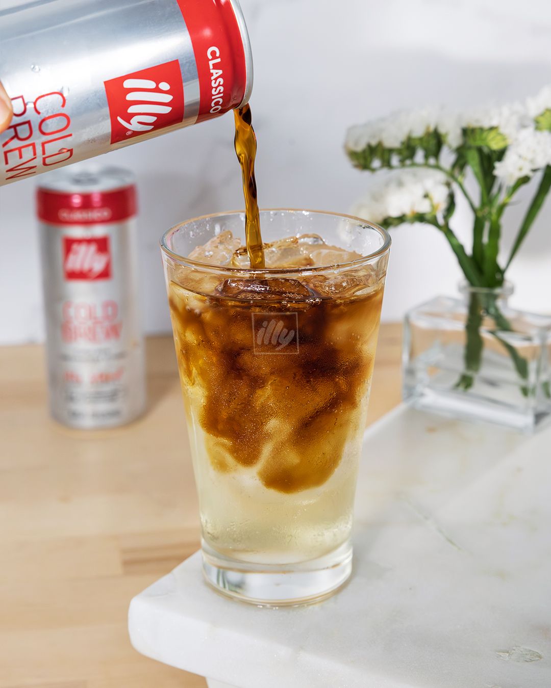 Cold brew coffee being poured over ice in glass with white flowers in background.