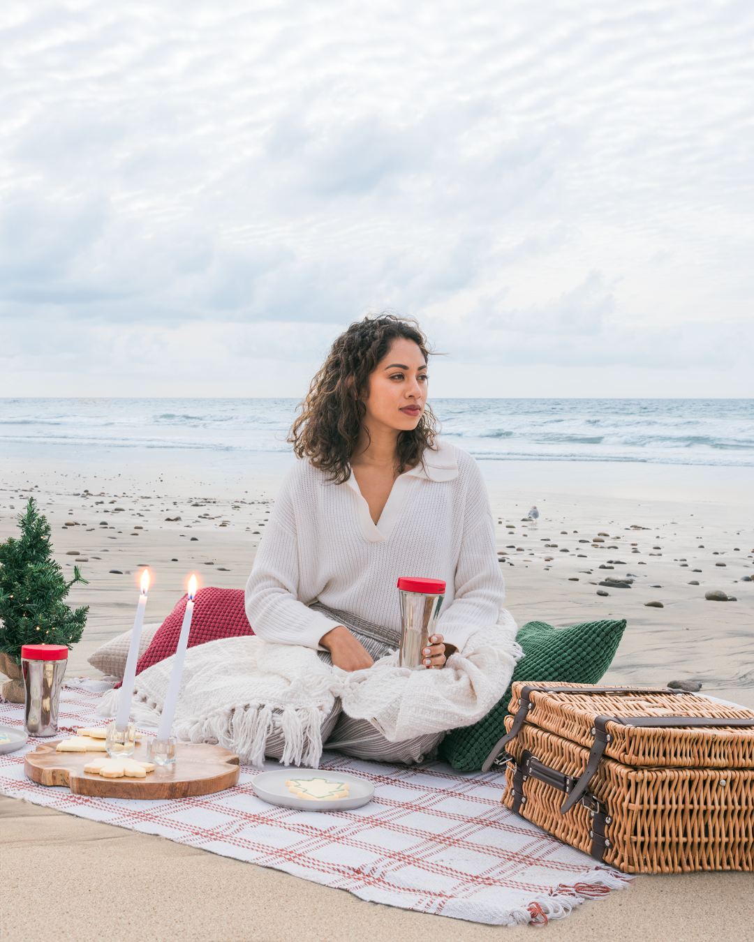 Cozy beach picnic setup with blanket, basket, and travel mug by the ocean.