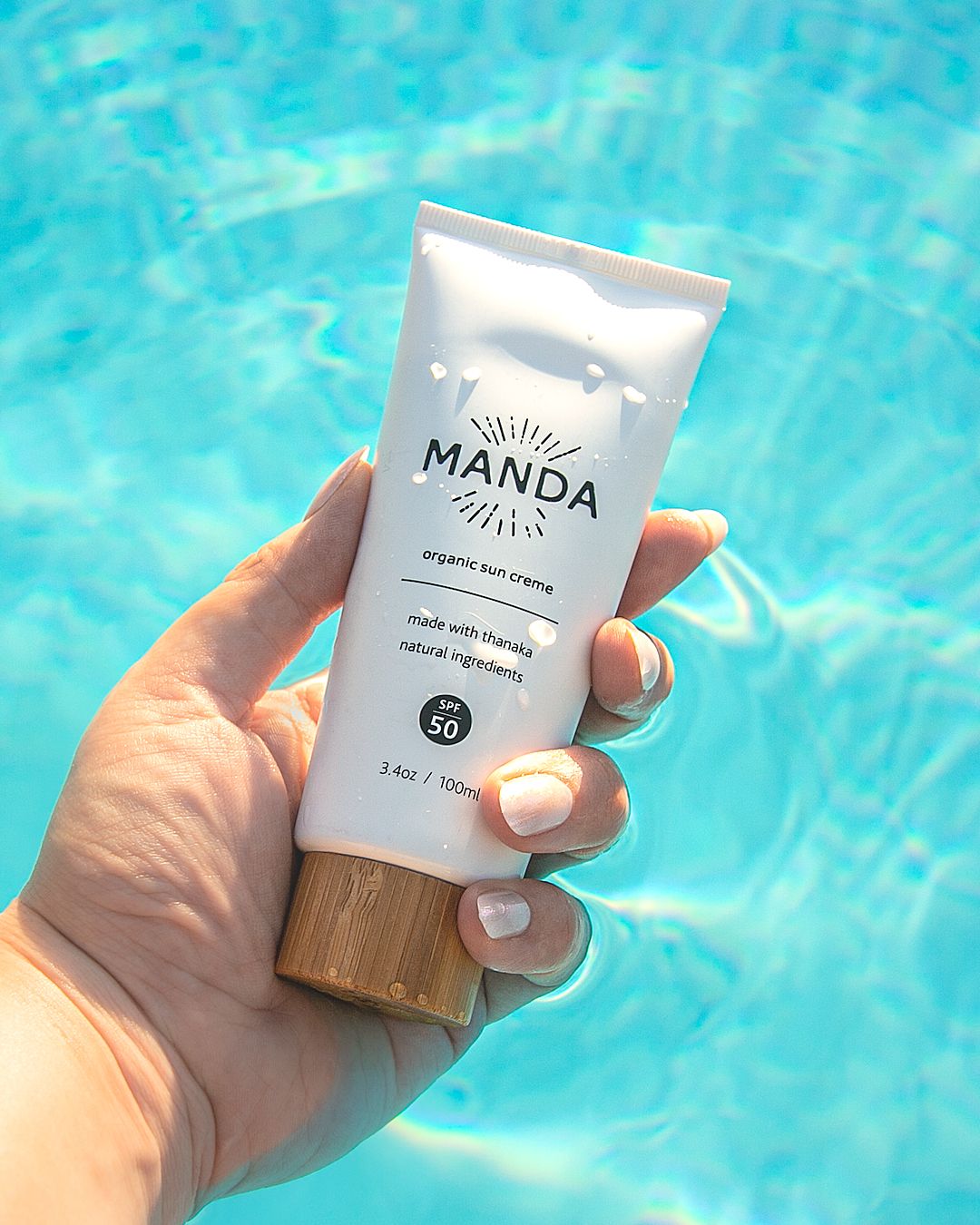 Hand holding white Manda skincare tube over sparkling turquoise pool water.