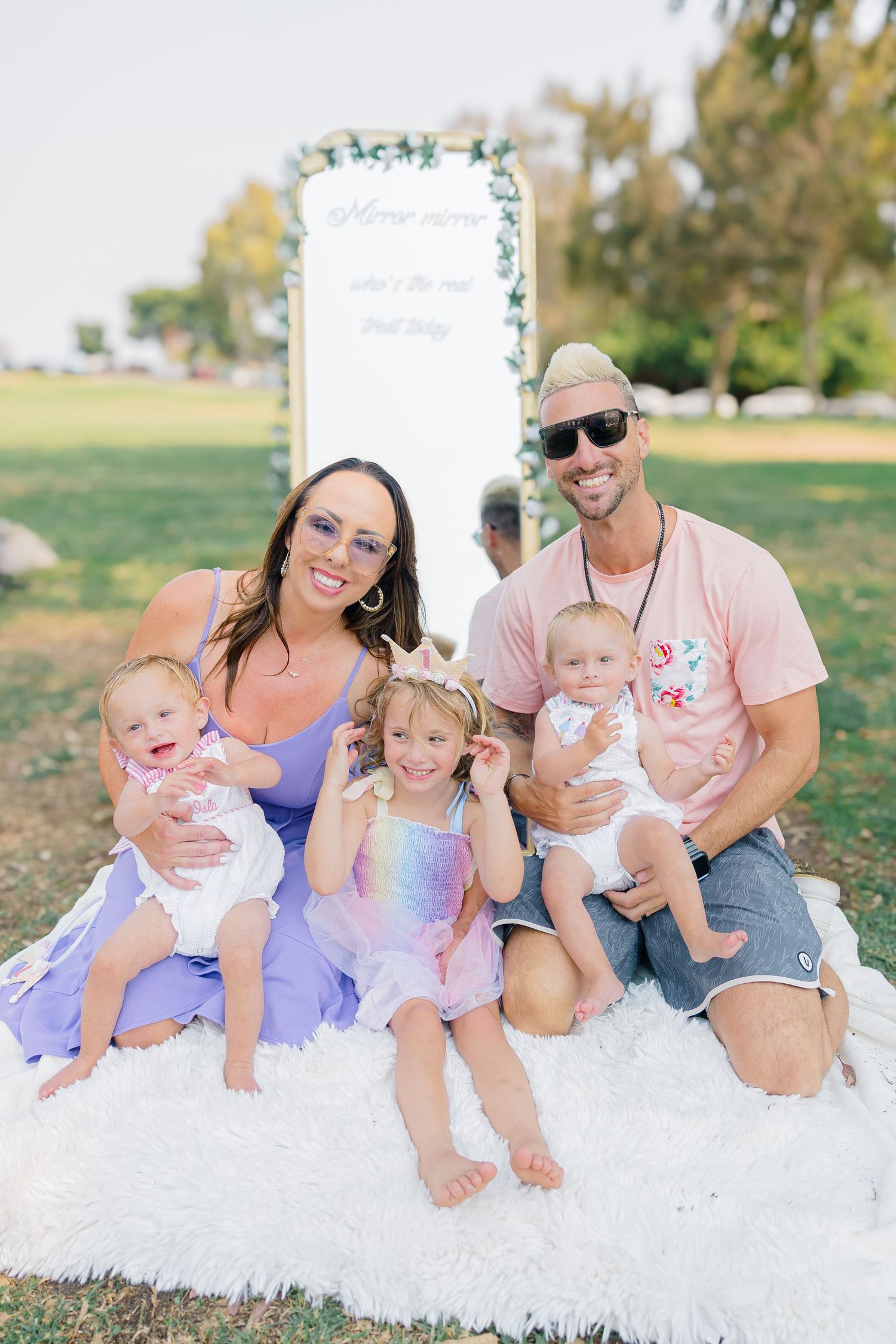 Family group seated on white blanket during outdoor photo session with pastel outfits.