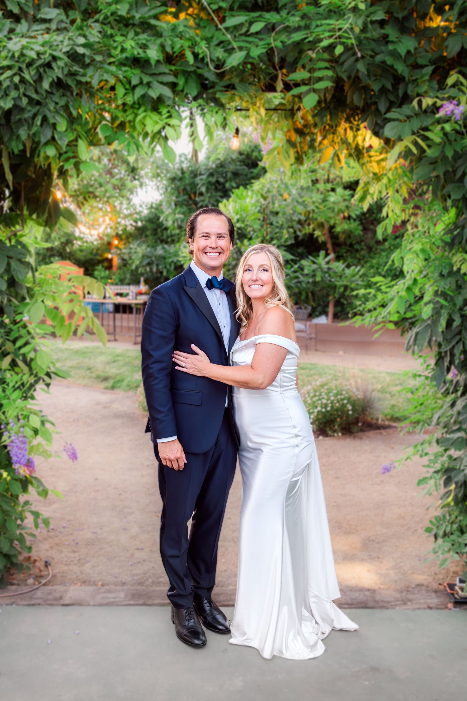 Wedding couple poses in lush garden setting with evening light filtering through trees.