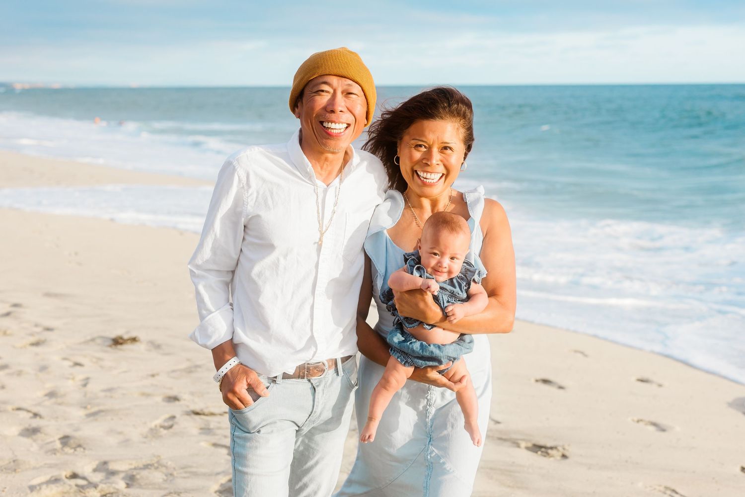 A family of three poses together on a sunny beach wearing coordinated white and blue outfits.