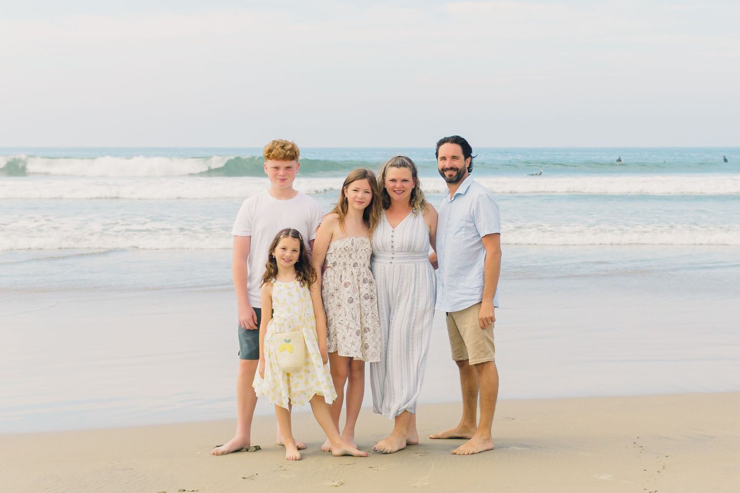 A family group stands together on a sandy beach at sunset wearing light casual beach attire.