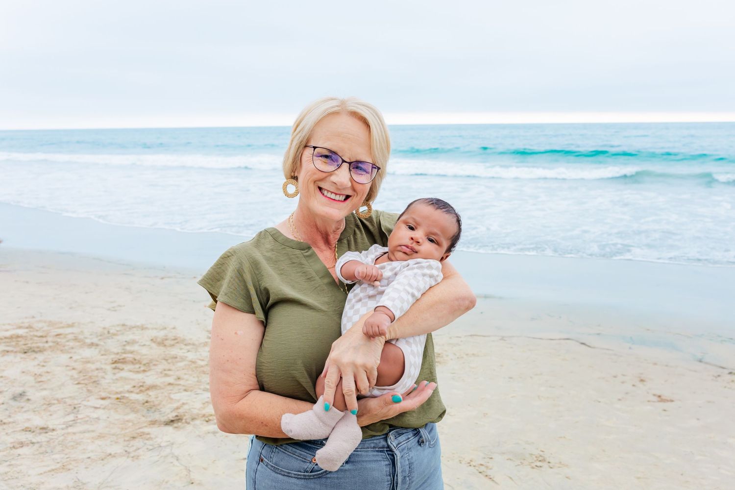 Adult holding young baby on beach with turquoise ocean waves in background.