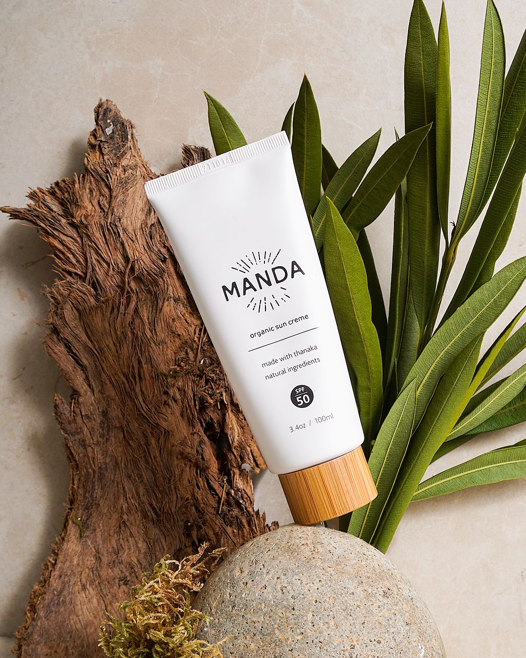 White and black Manda skincare tubes displayed with tropical leaves and natural wood decor elements.