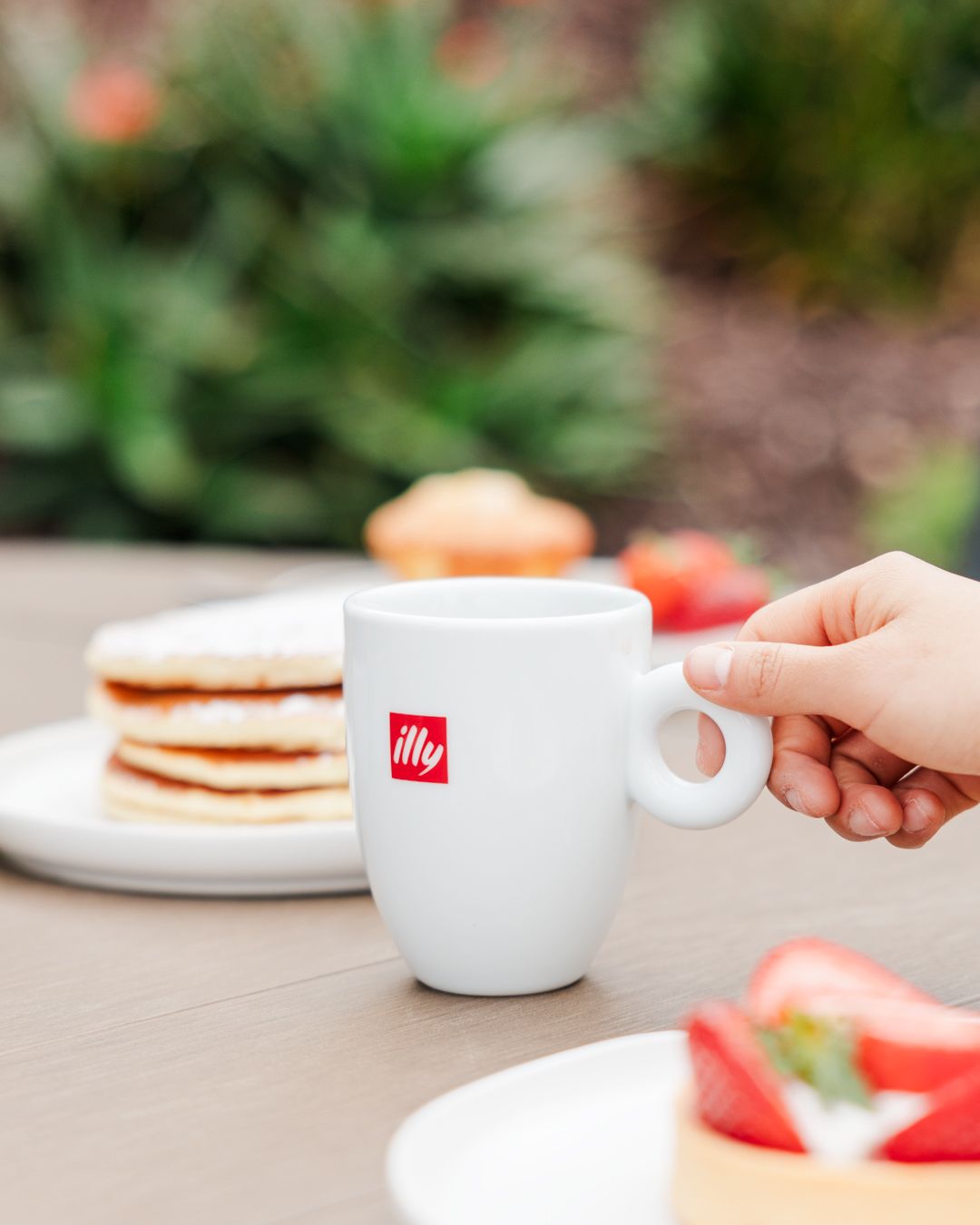 White illy coffee cup with stack of pancakes and fresh strawberries on outdoor table.