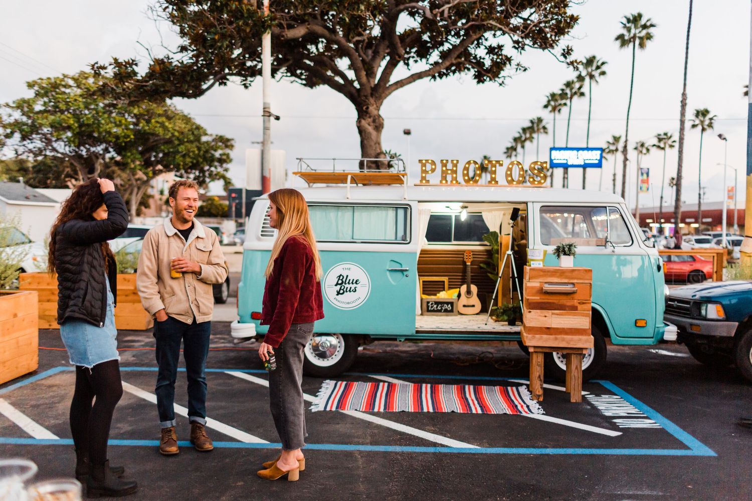 Group of people chatting near vintage blue VW vans in a beachside parking lot with palm trees.