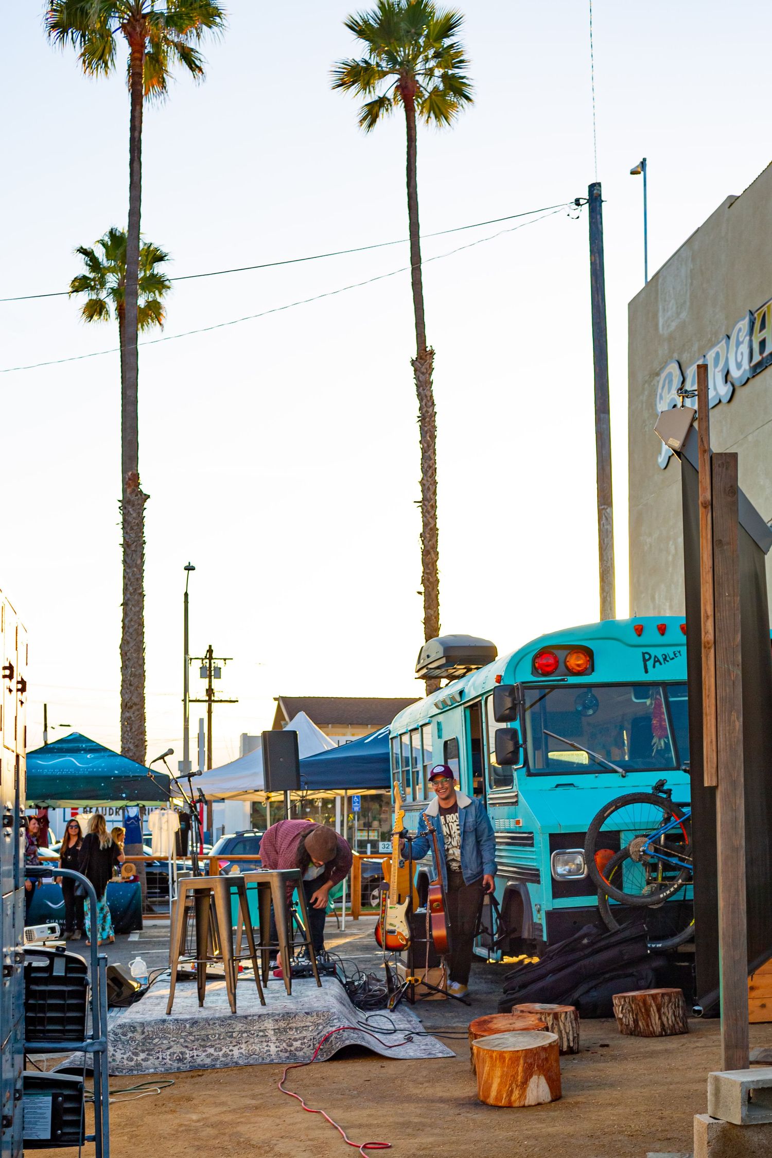 Row of teal-colored vendor tents and palm trees line a sunny outdoor market area at sunset.
