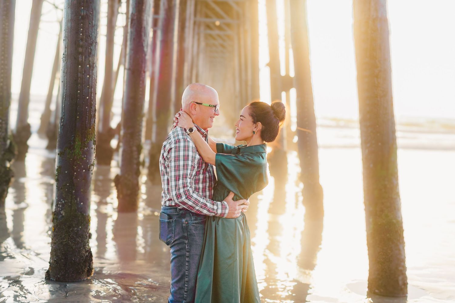 A couple embraces under a pier at sunset with golden light streaming through the wooden pillars.